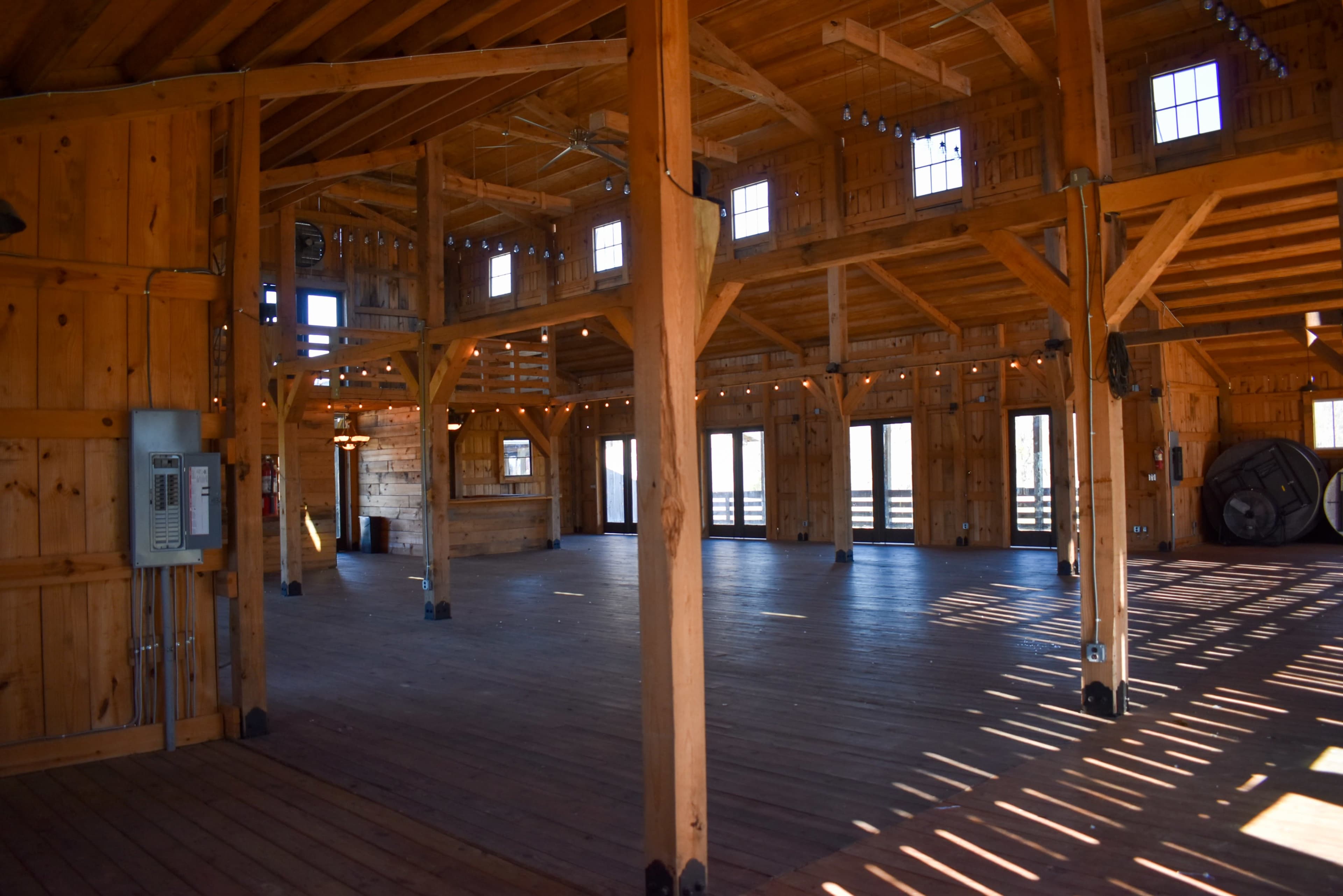 The image shows the interior of a wooden barn with exposed beams and a spacious wooden floor, illuminated by natural light from large windows.