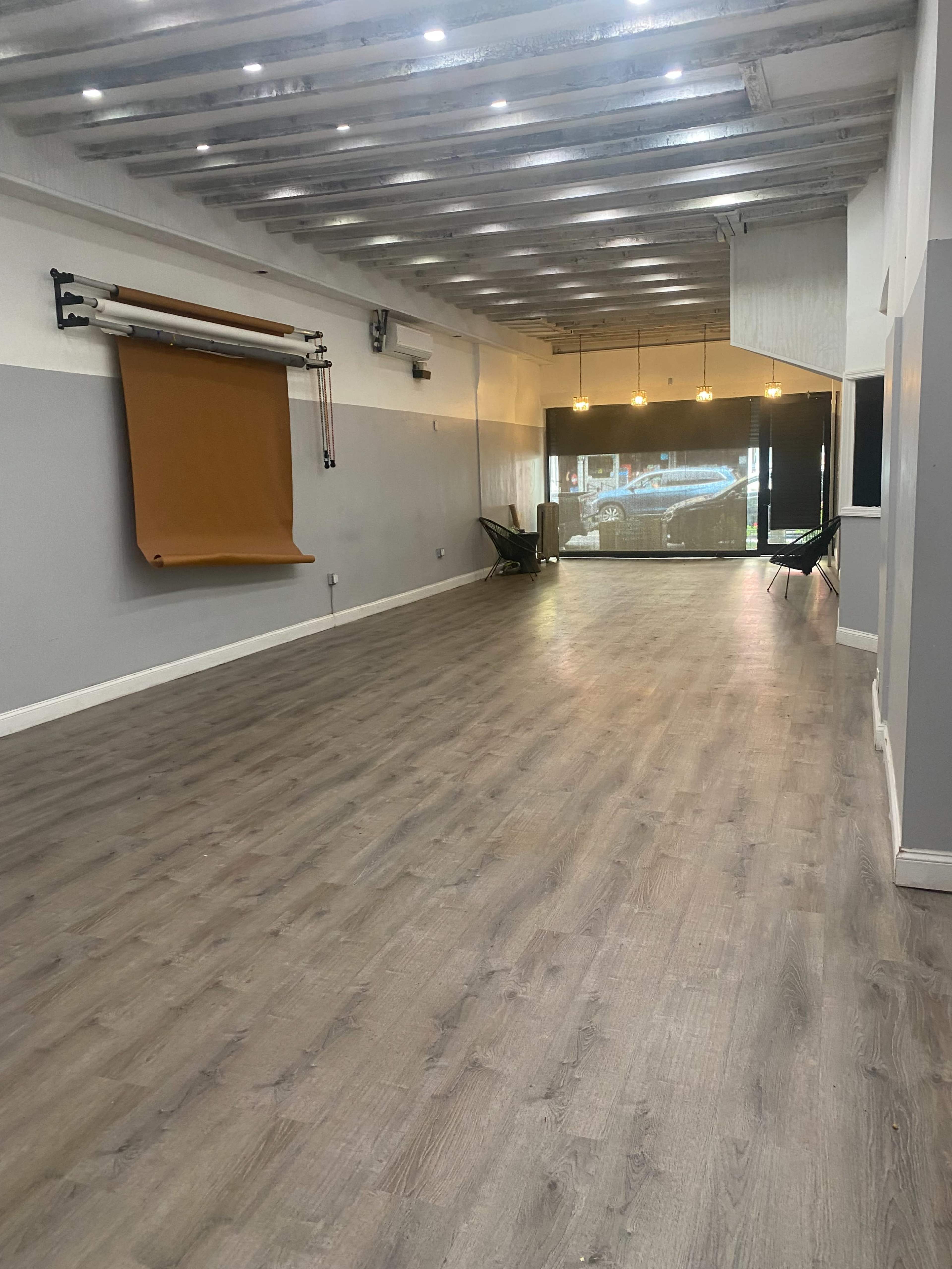 An empty room with wooden flooring, a brown backdrop on the wall, and large windows allowing natural light.
