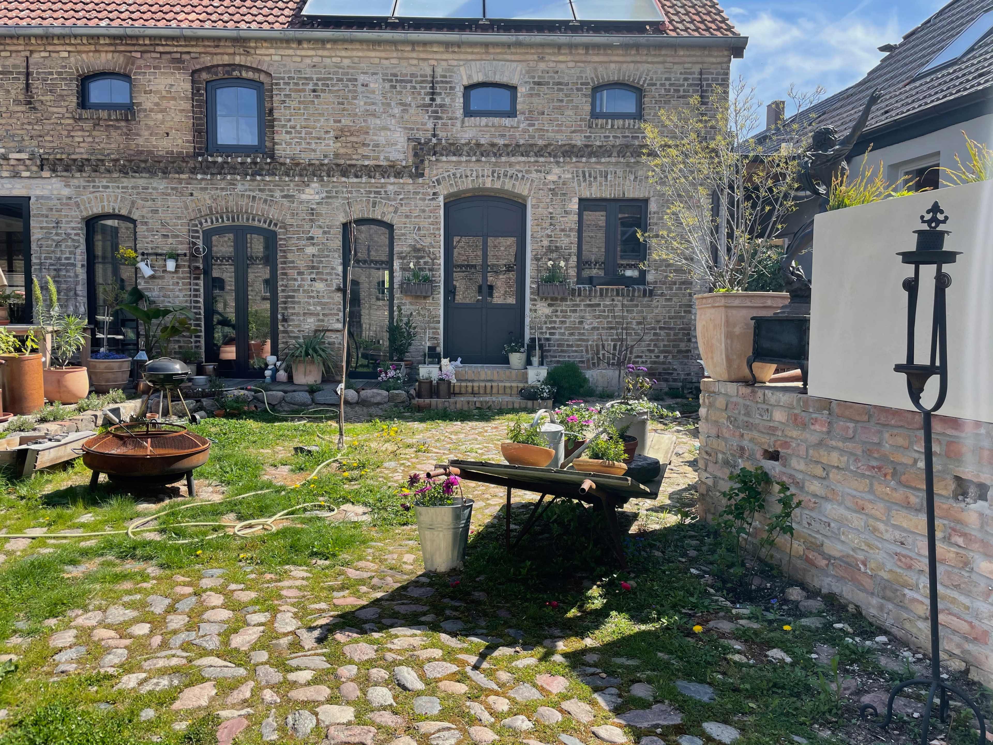 The image shows a cobblestone courtyard with a brick building featuring arched windows and a door, surrounded by various potted plants and a table with gardening supplies.