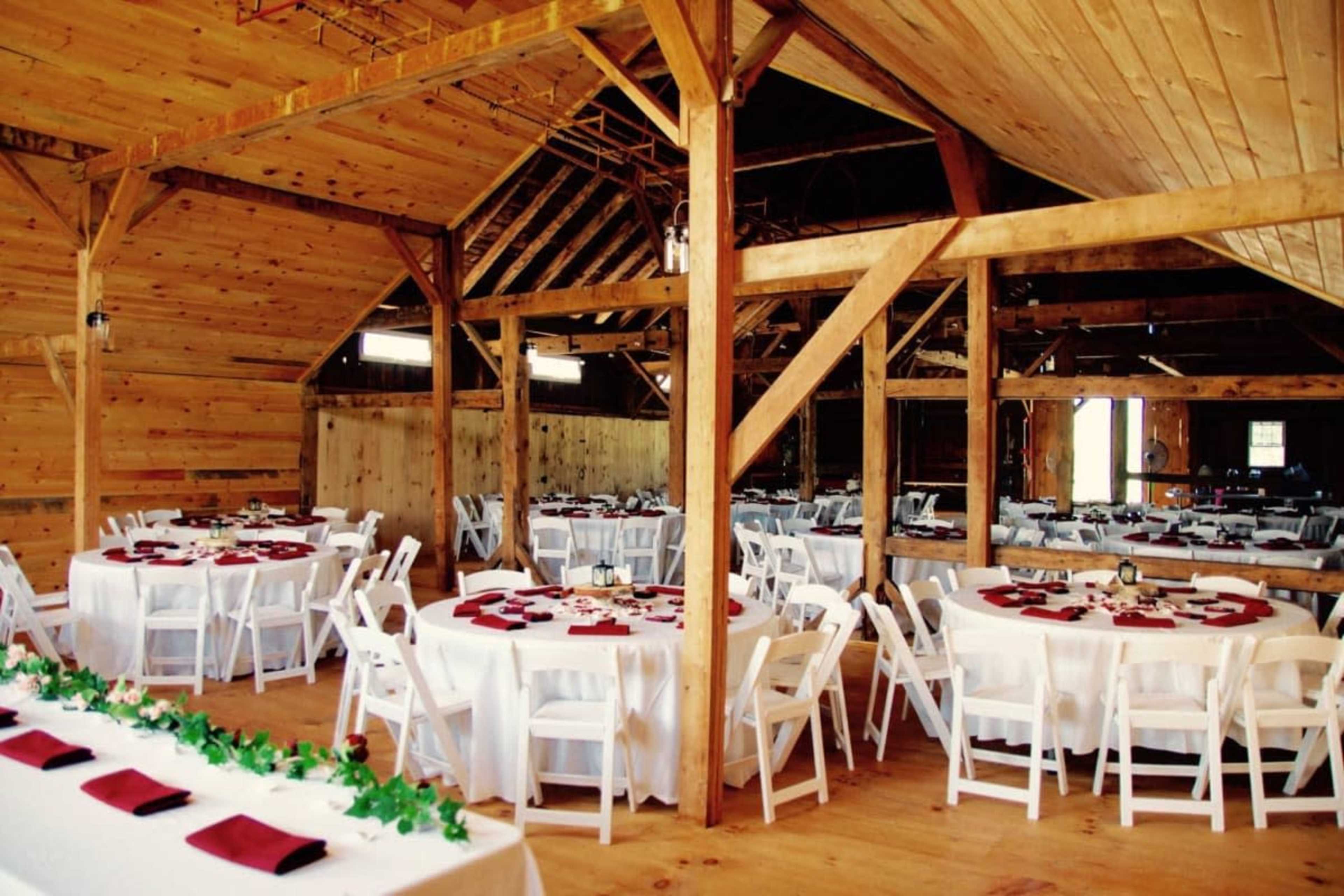 The interior of a wooden barn is set up for a dinner event, featuring round tables covered with white tablecloths and adorned with maroon napkins.