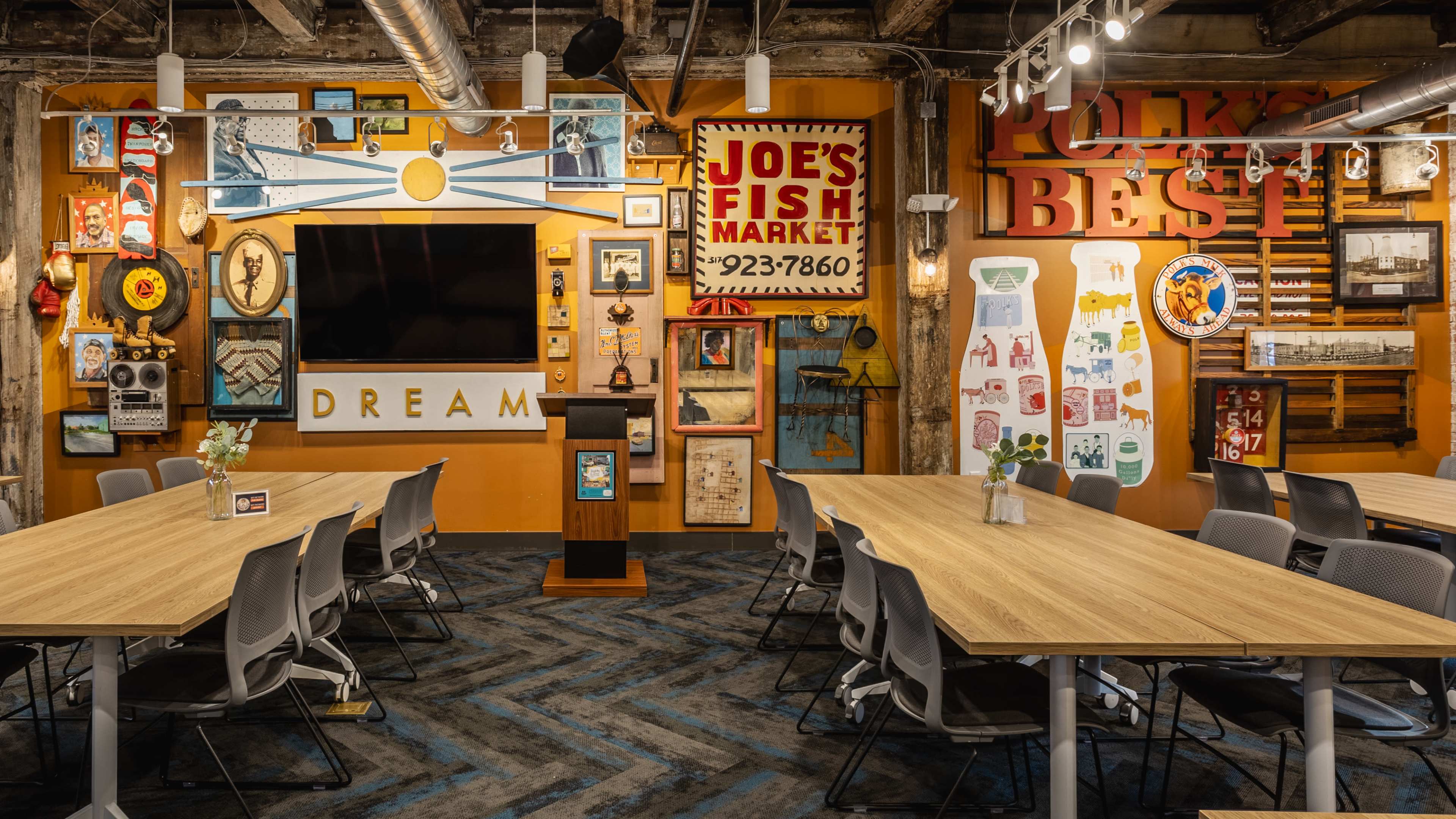 The image shows a modern meeting room with long tables and chairs arranged in front of a wall adorned with various vintage signs and artwork.