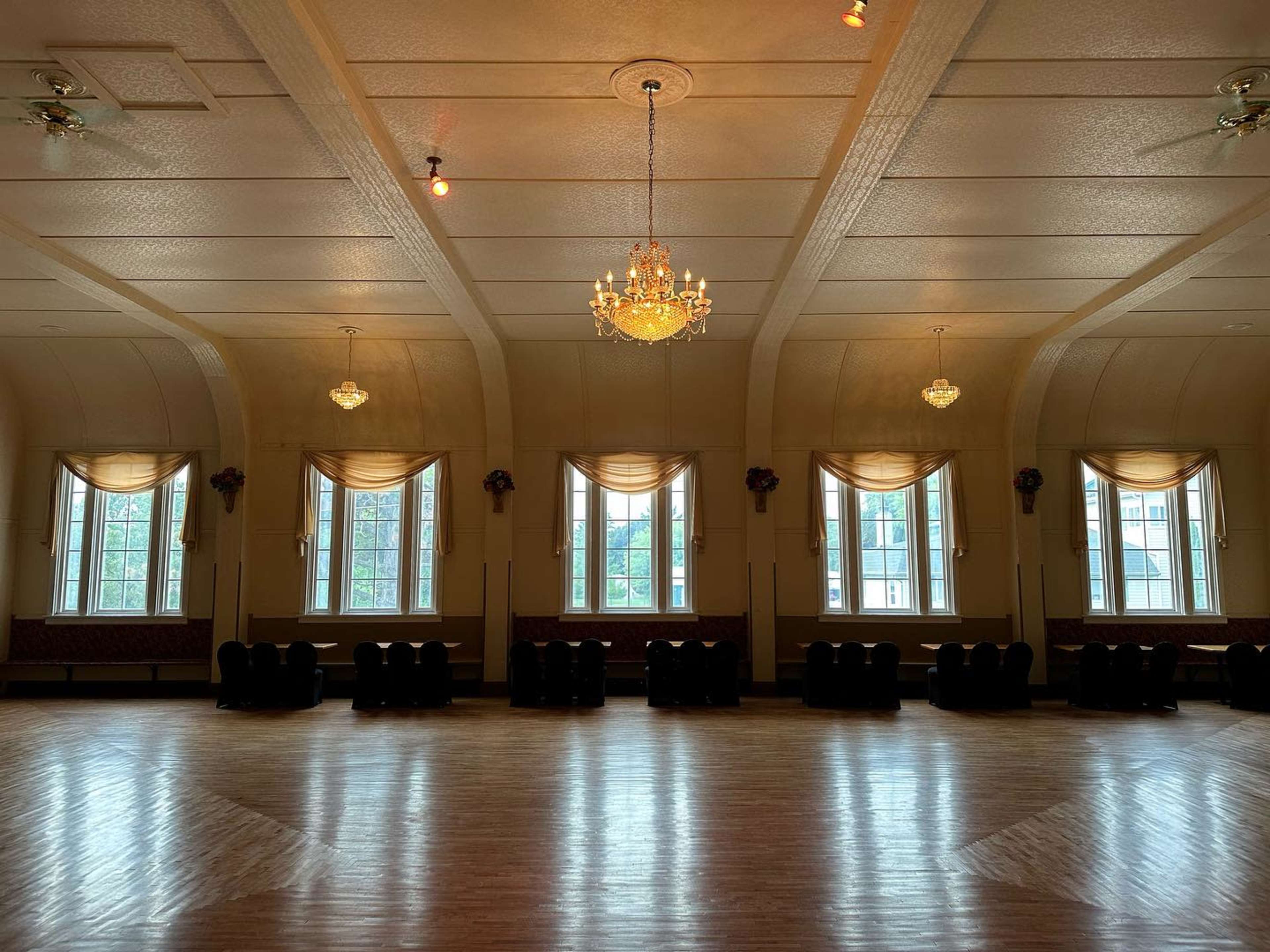 A large, empty hall with wooden flooring, adorned with a chandelier and multiple windows dressed with draped curtains.
