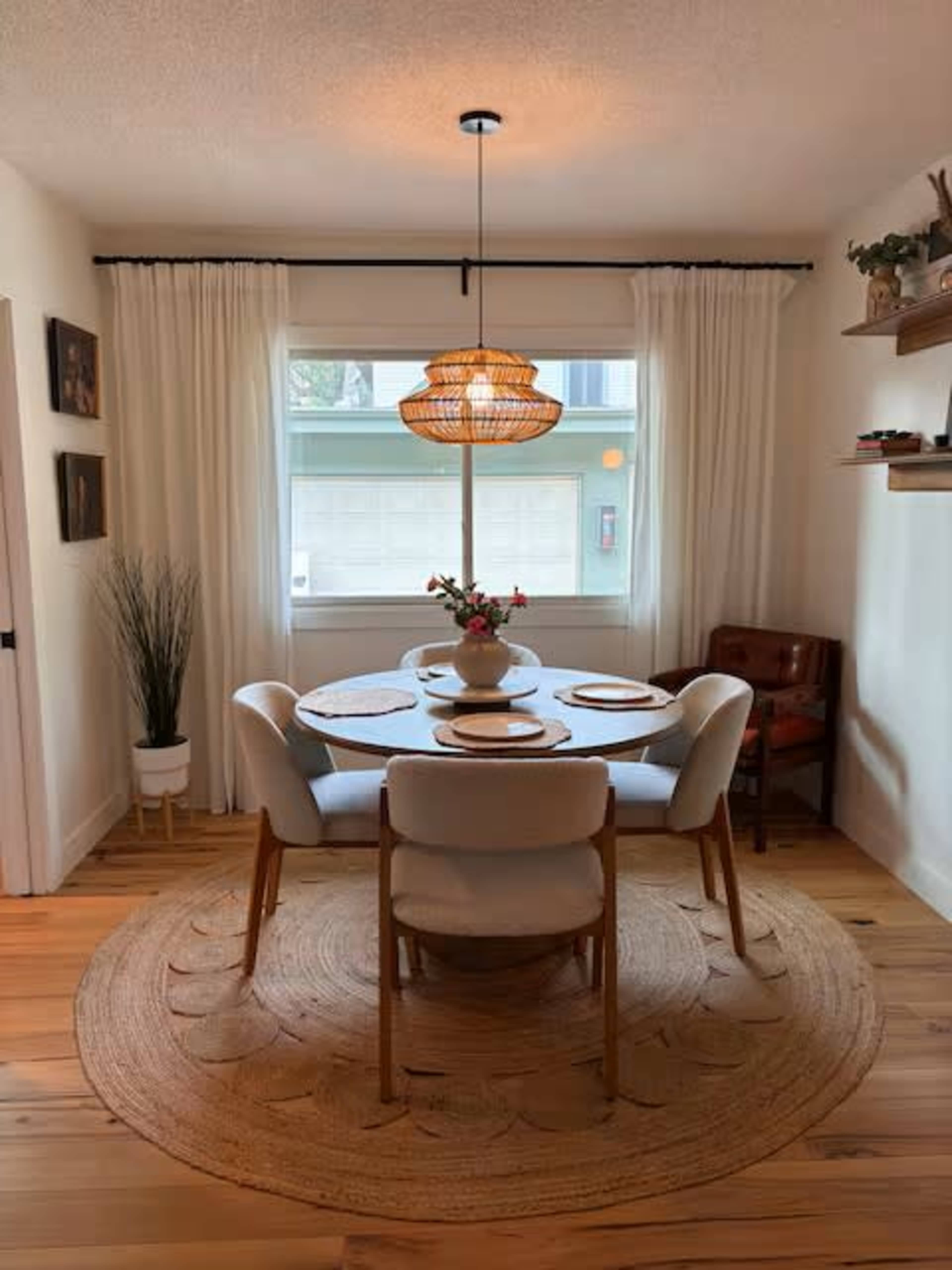 A circular dining table is set with plates and a vase, surrounded by four chairs, in a room with a large window covered by white curtains.