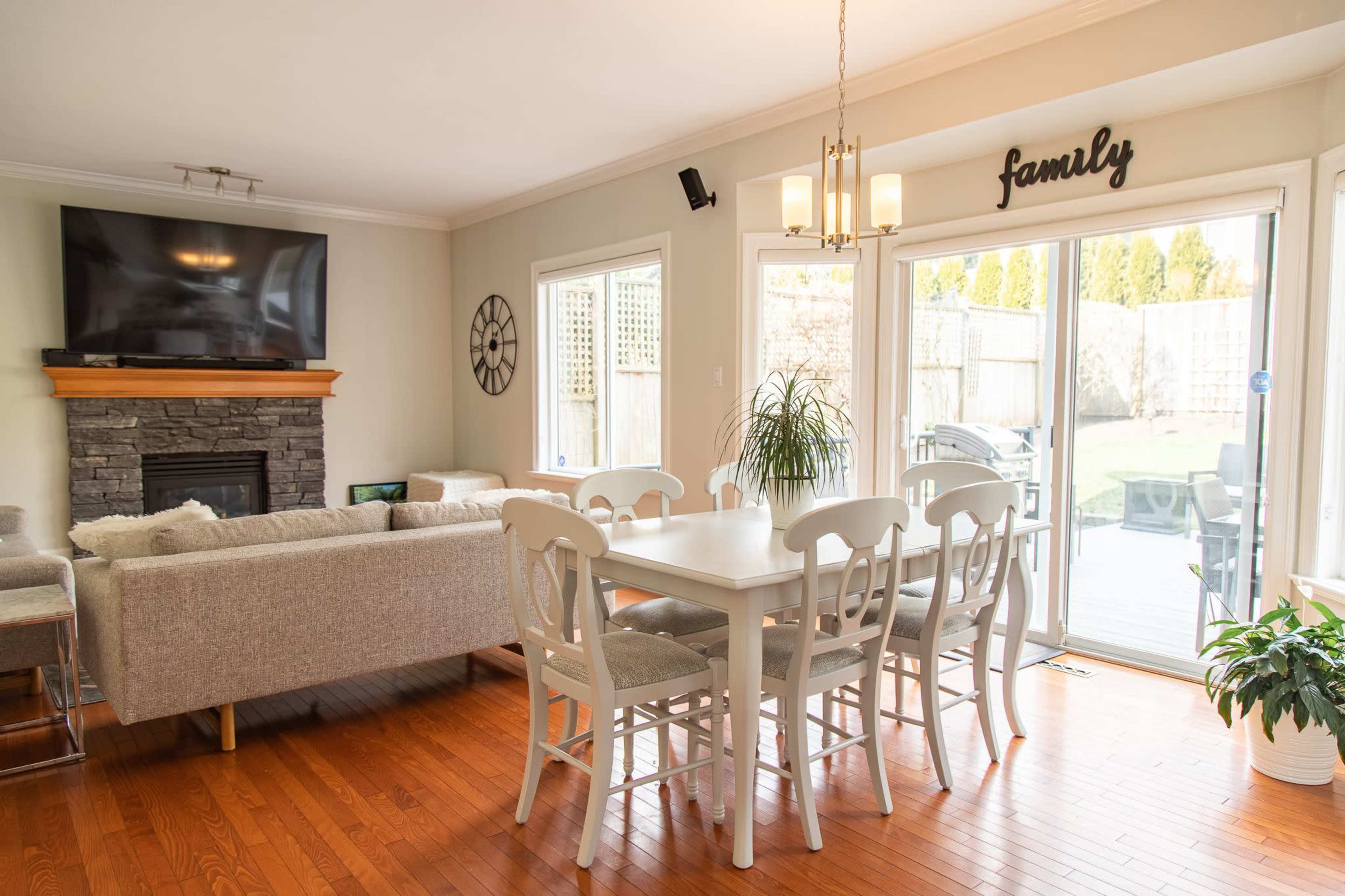 A bright living and dining area with a wooden dining table and chairs, a stone fireplace, and large windows overlooking a backyard.