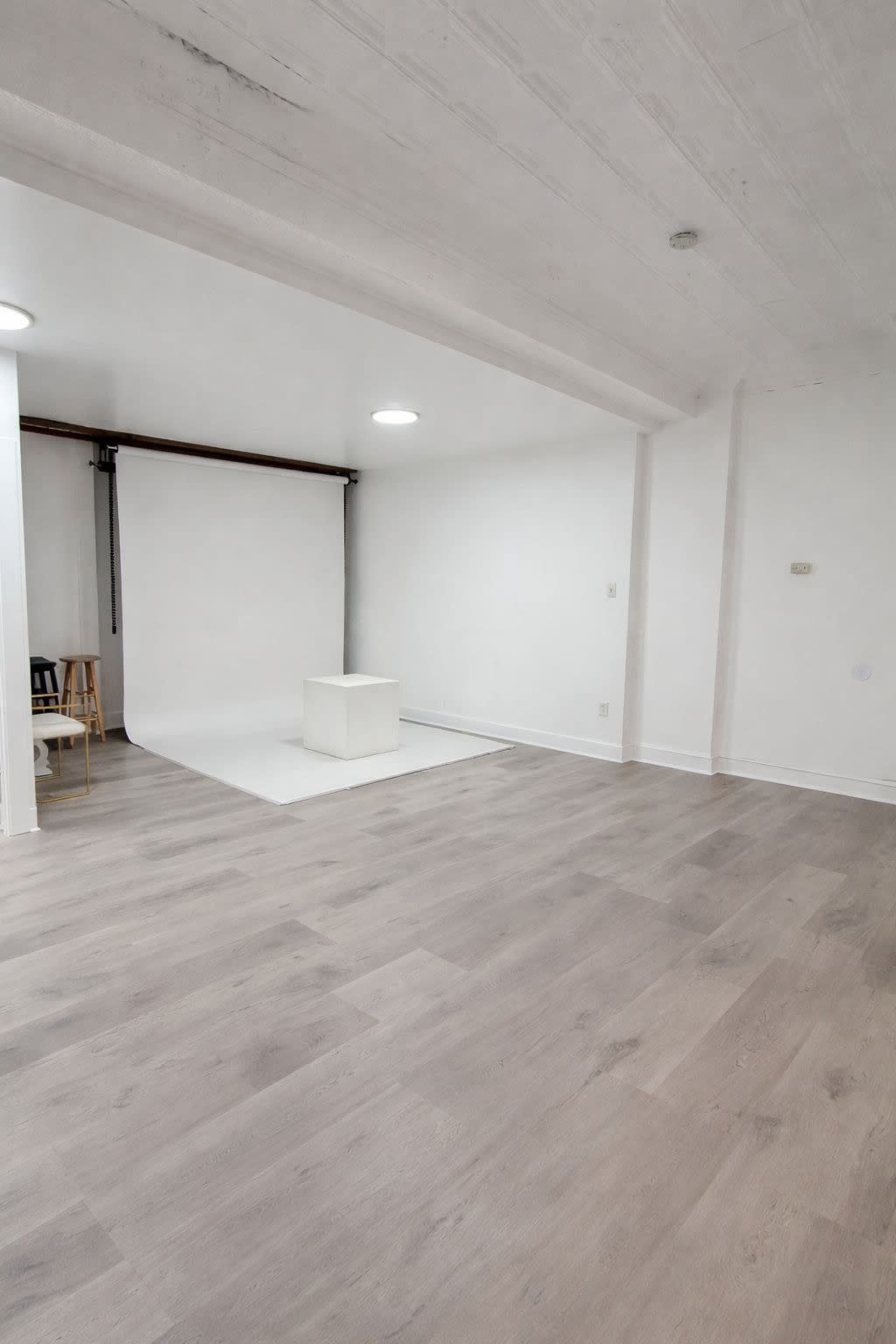 The image shows a spacious, empty room with light-colored wooden flooring and a simple white backdrop, featuring a cube in the center and a chair in the corner.