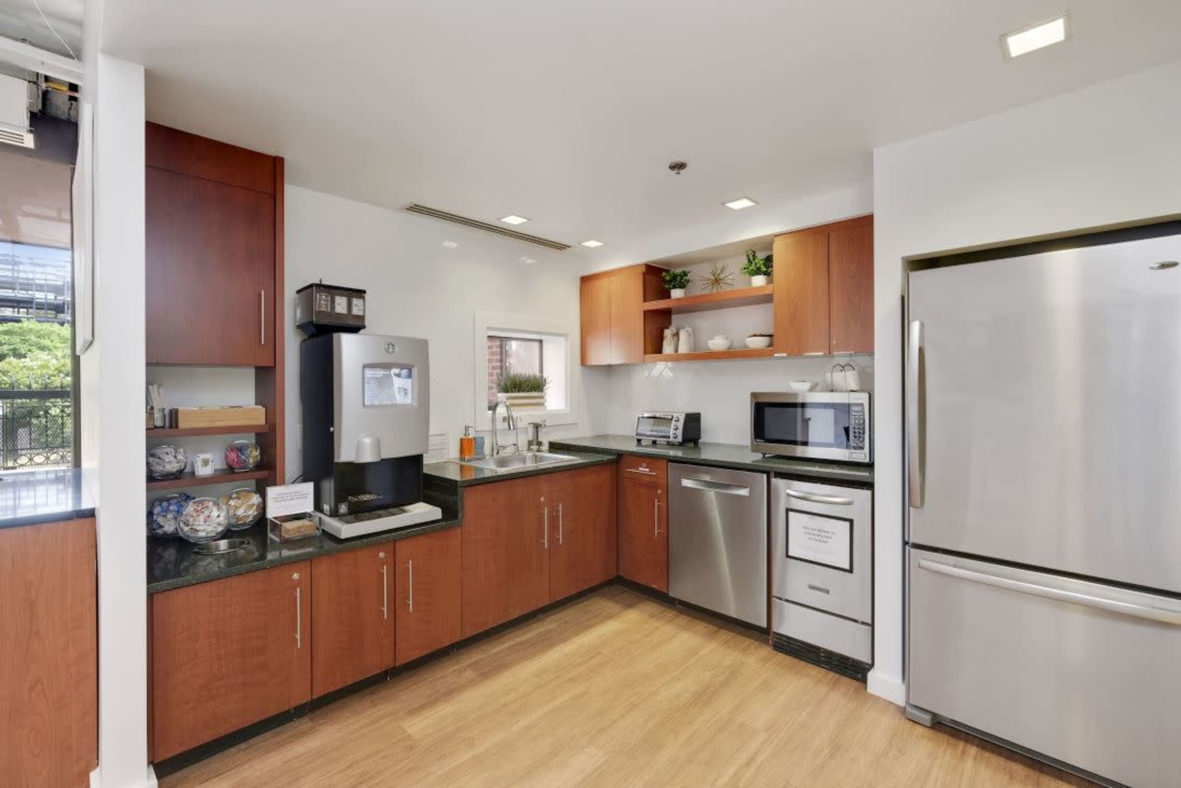 The image shows a modern kitchen featuring wooden cabinets, stainless steel appliances, and a dark countertop with various kitchen accessories.