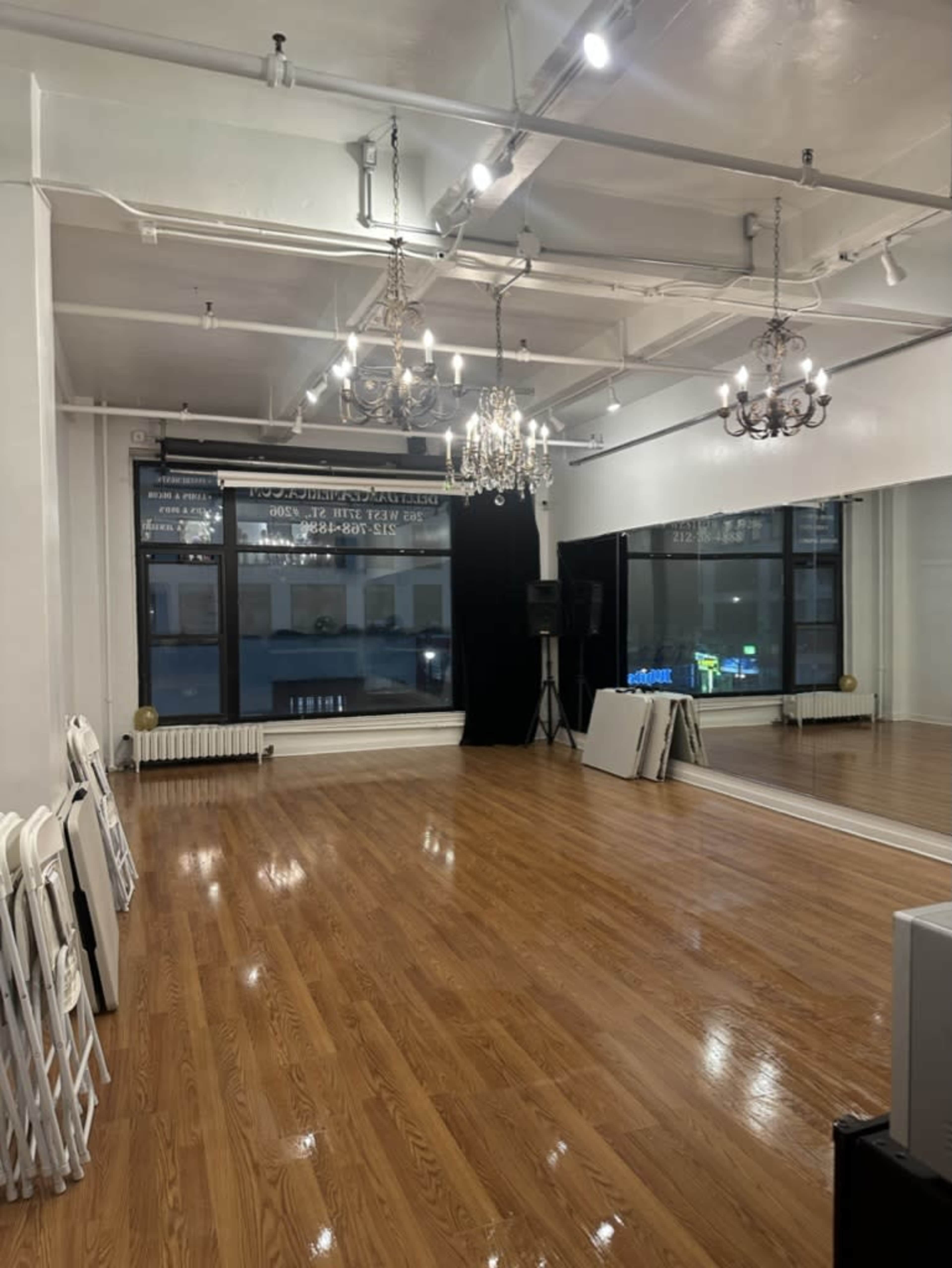 The image shows an empty dance studio with polished wooden floors, white walls, and large windows, illuminated by multiple light fixtures.