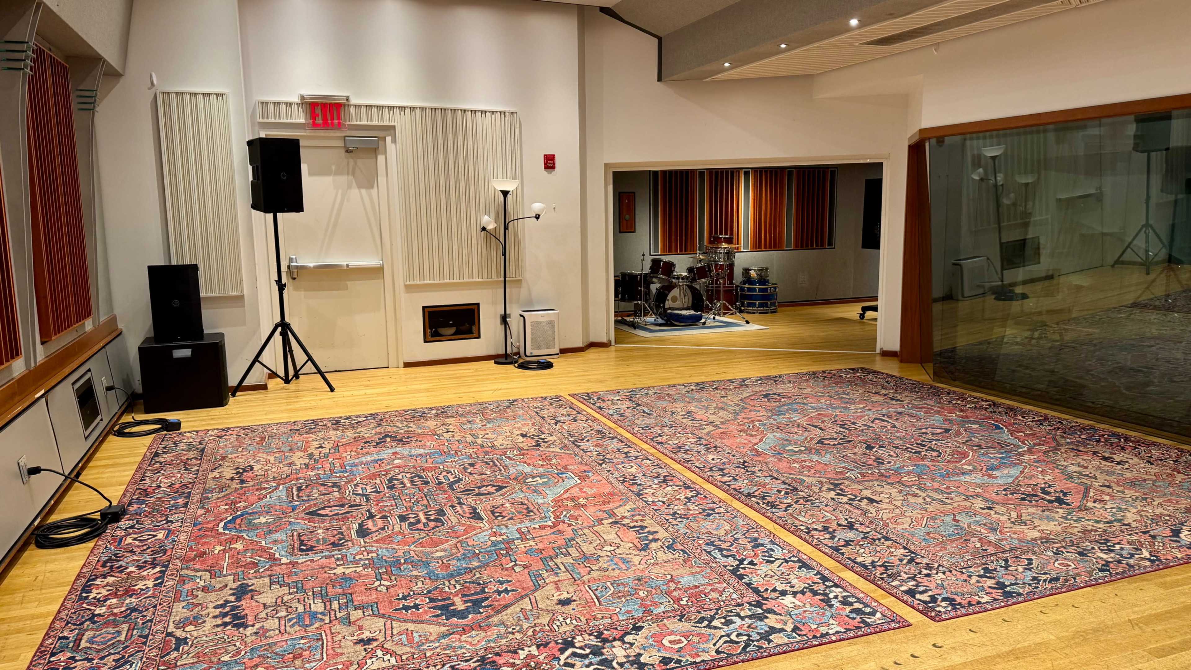 The image shows a spacious music studio with wooden floors, large rugs, and a glass wall separating the control room from the recording area.