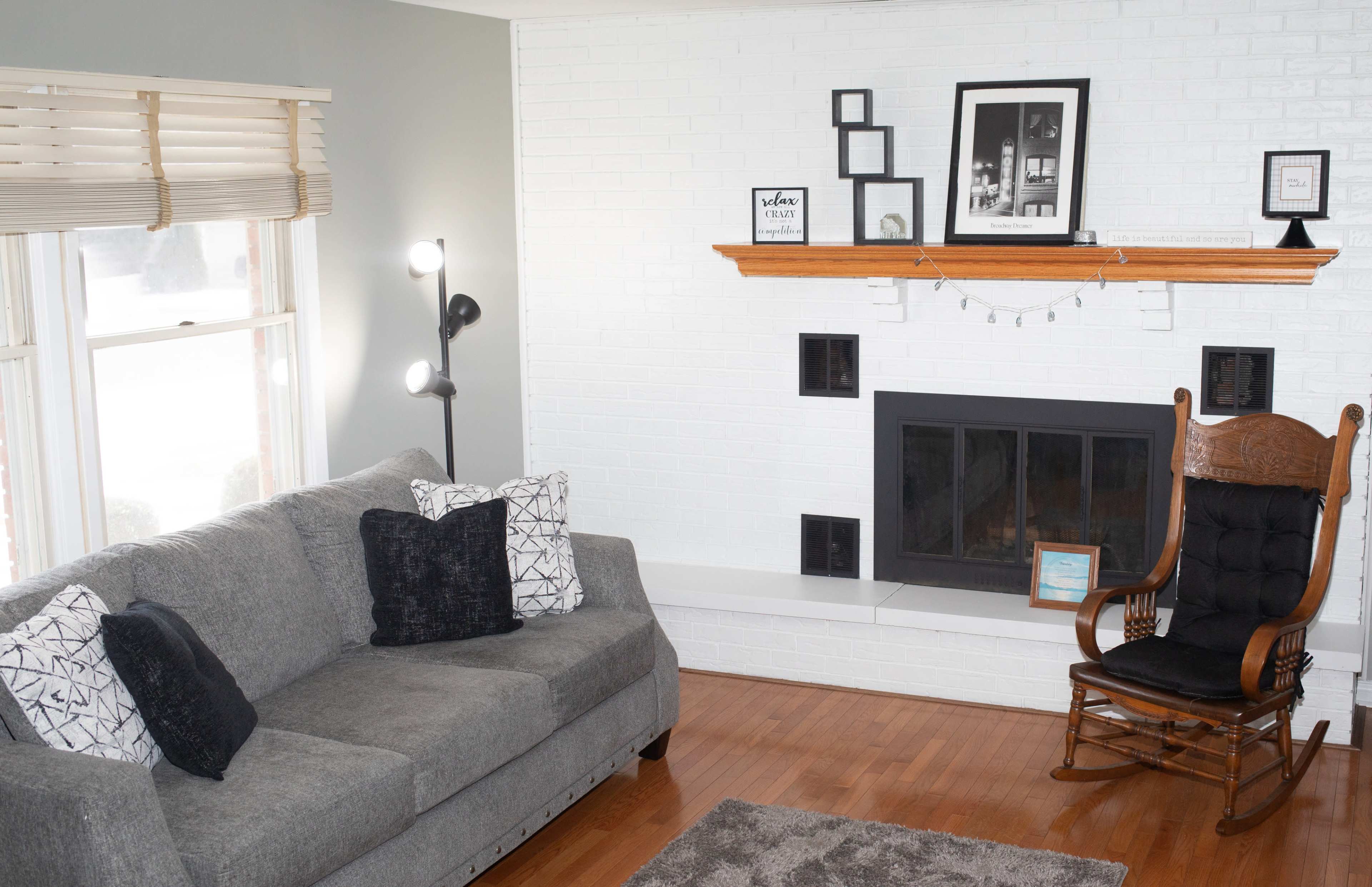 The living room features a gray sofa with decorative pillows, a wooden rocking chair, and a white brick fireplace with a wooden mantel displaying framed photos.