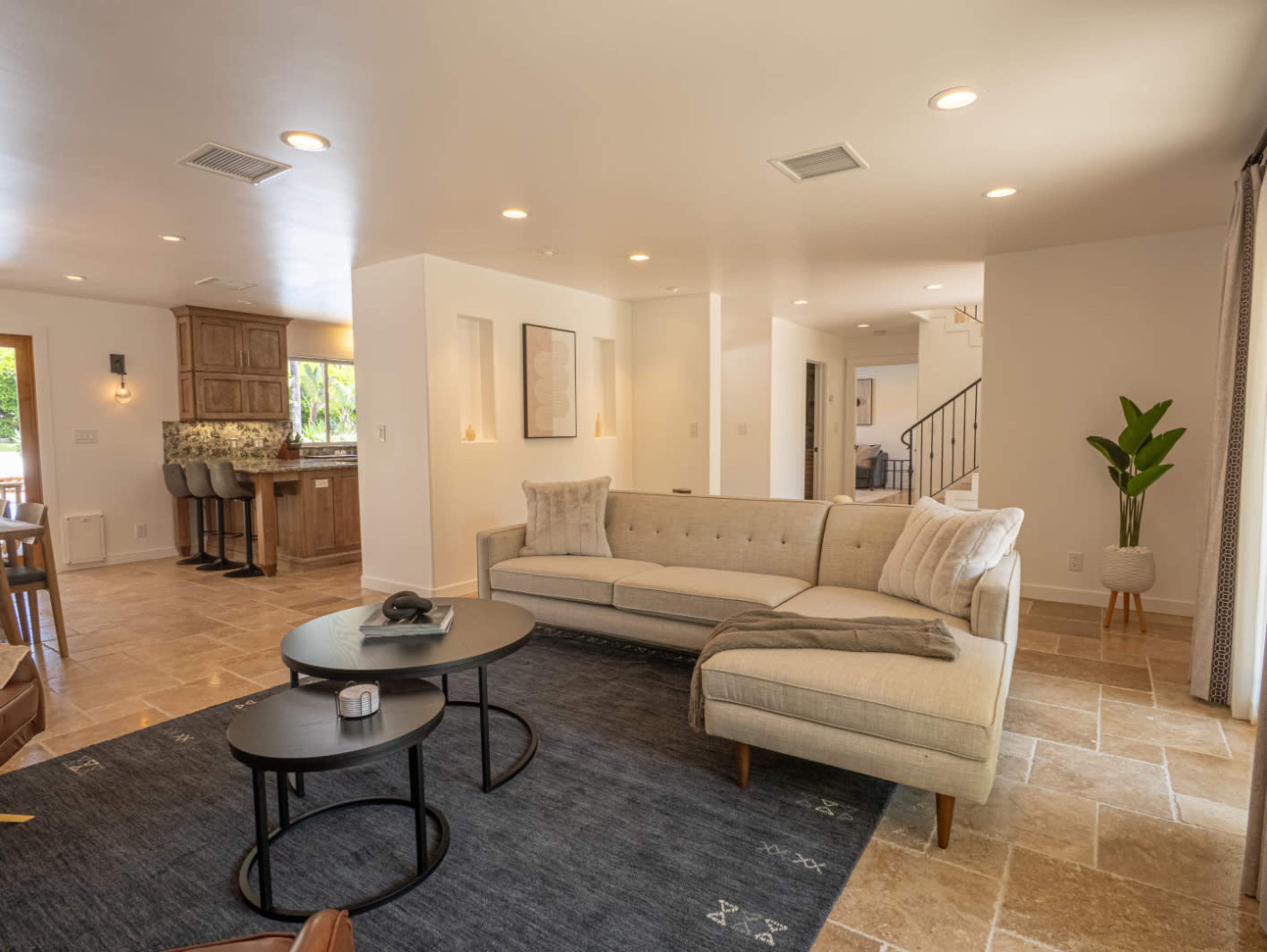 A modern living room featuring a light-colored sectional sofa, a round coffee table, and natural stone flooring, with a kitchenette visible in the background.