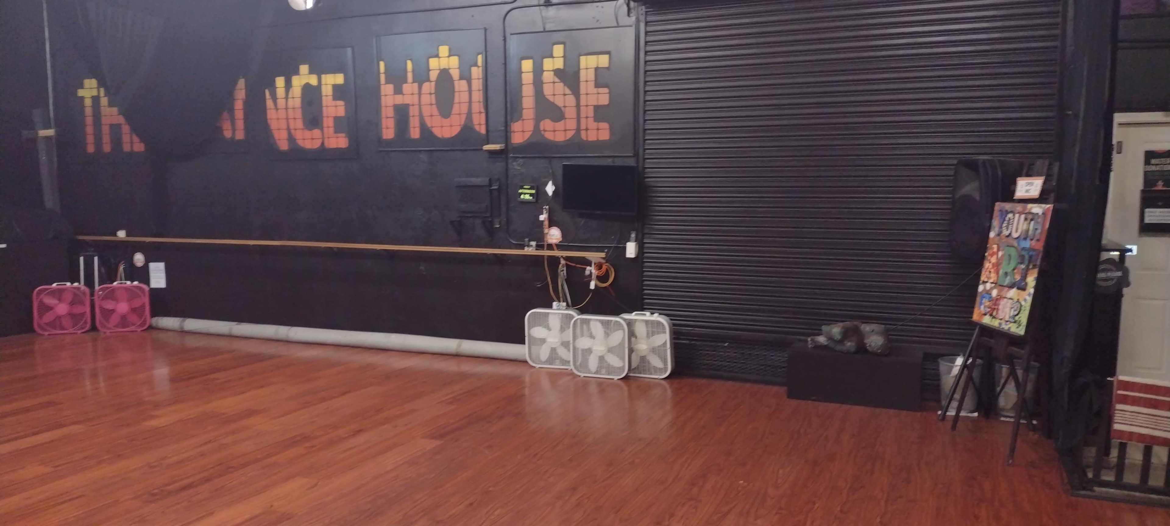 A dance studio with hardwood flooring, a wall featuring the words "THE DANCE HOUSE," and equipment including fans and a television on a black backdrop.