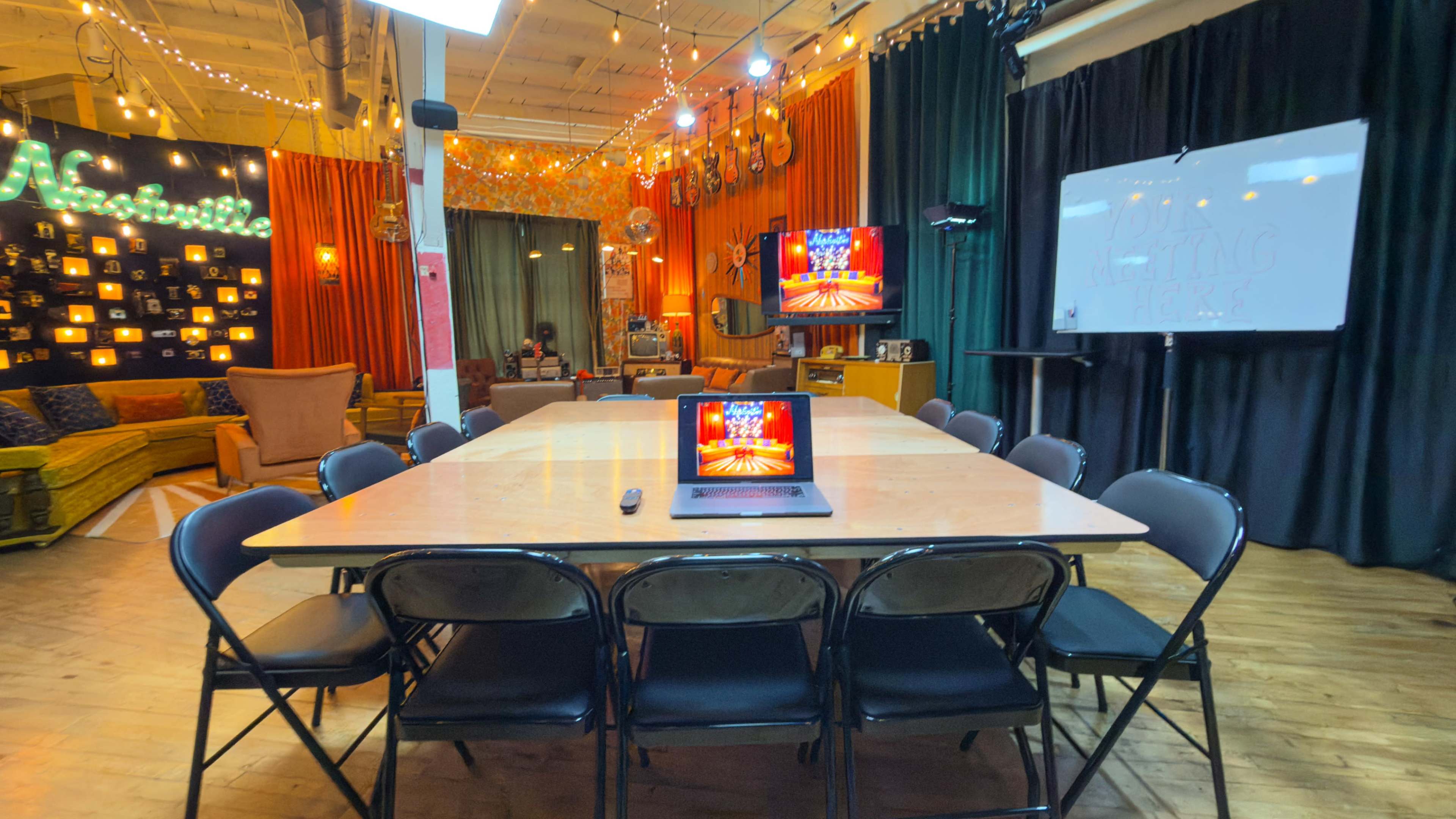 A long table surrounded by black chairs is set up in a colorful room decorated with string lights and screens displaying images.