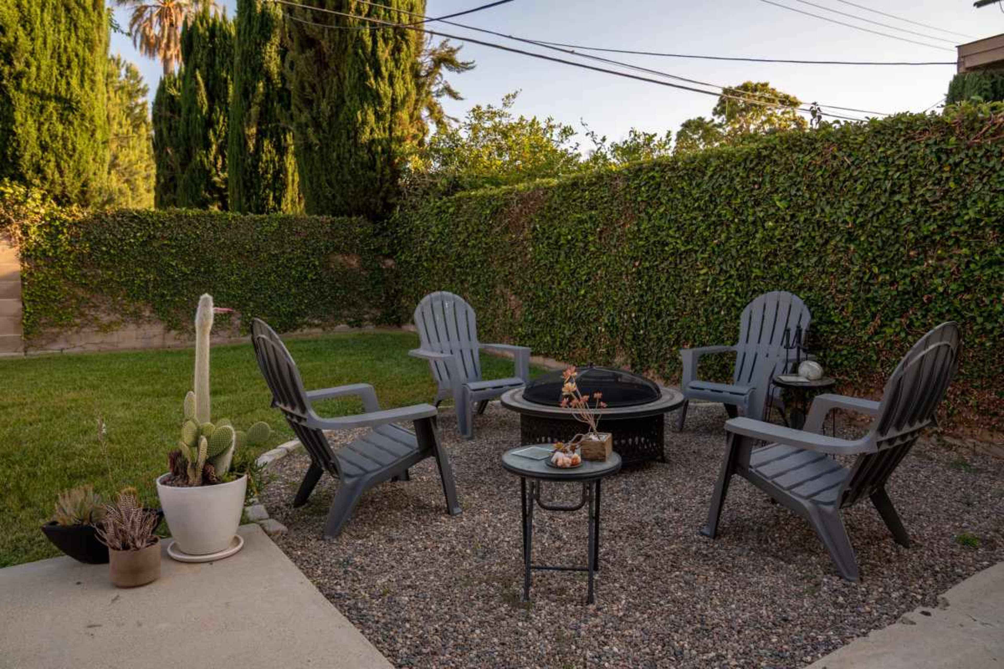A seated area in a garden features four gray Adirondack chairs arranged around a small fire pit, surrounded by a gravel surface and greenery.