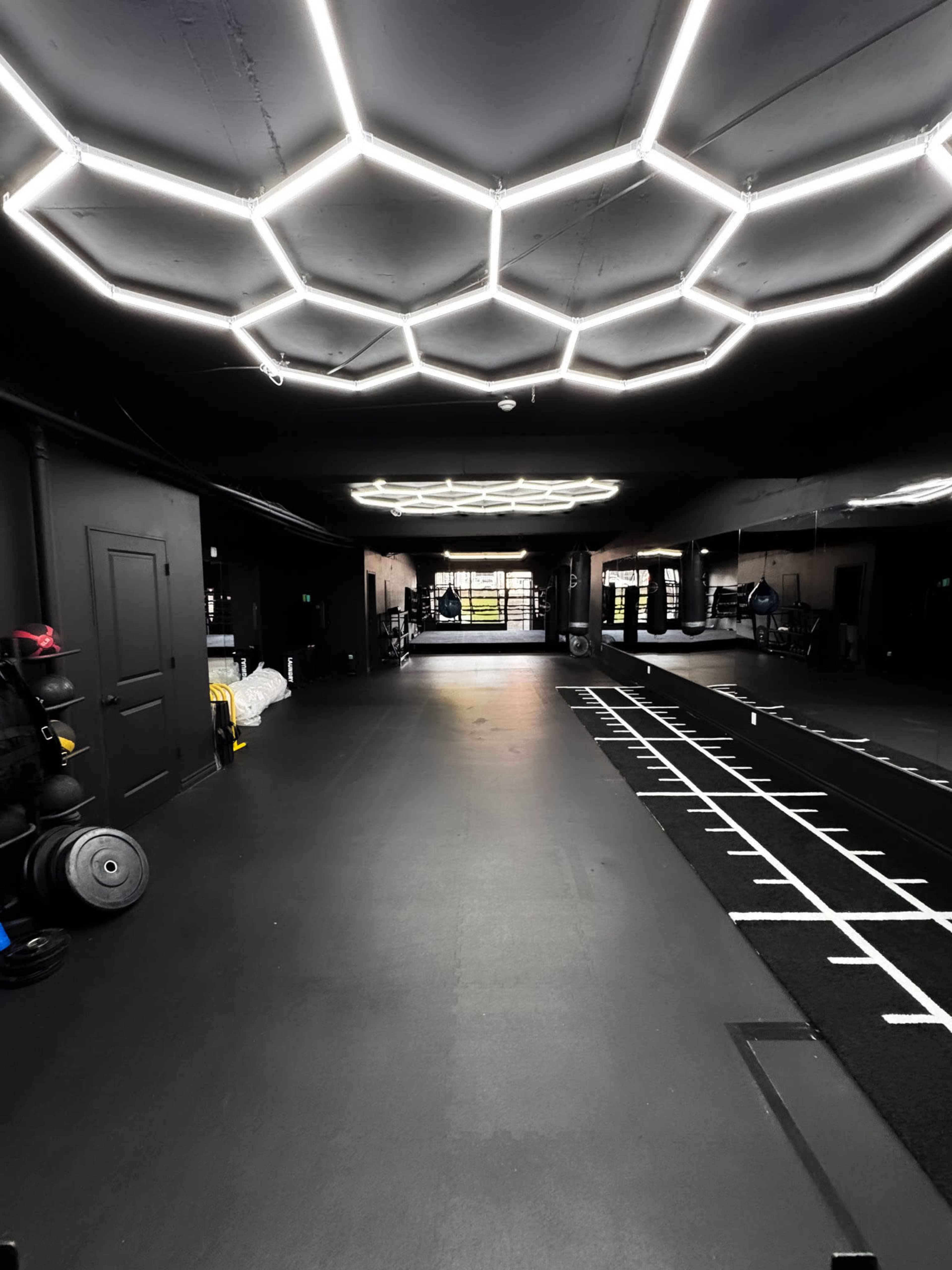 The image shows a modern gym interior with a hexagonal light fixture, black walls, and various fitness equipment arranged along the walls and floor.