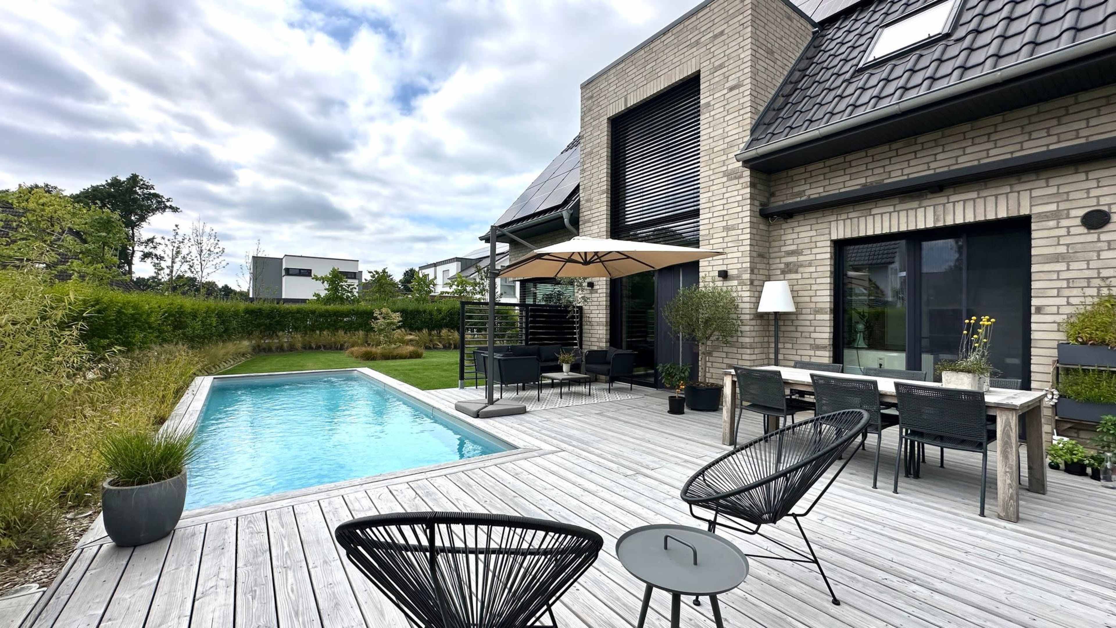 The image shows a modern backyard featuring a swimming pool, wooden deck, outdoor seating area, and well-maintained landscaping.