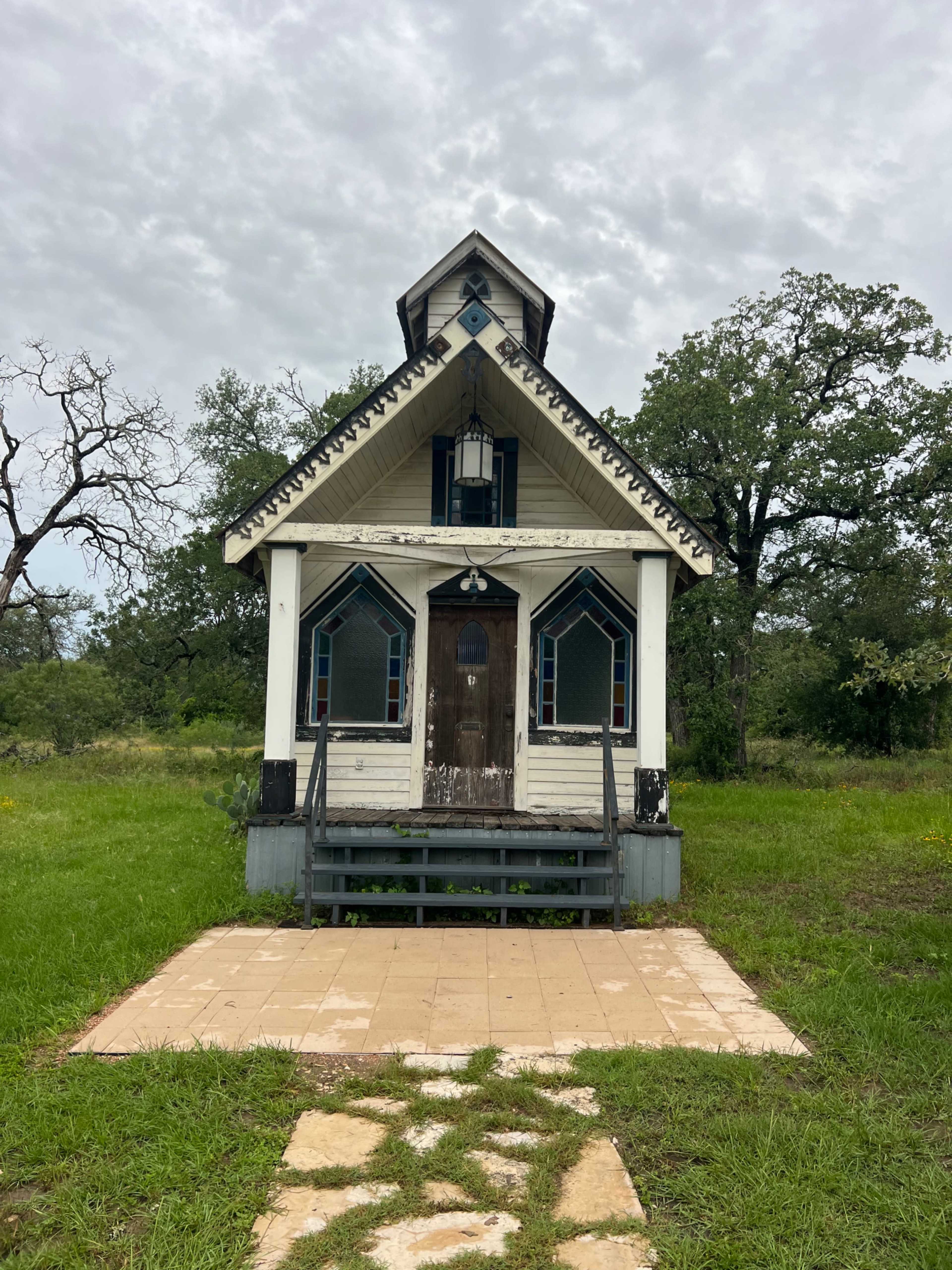 The image shows an old, weathered house with a steep roof, a front porch, and surrounding greenery under a cloudy sky.