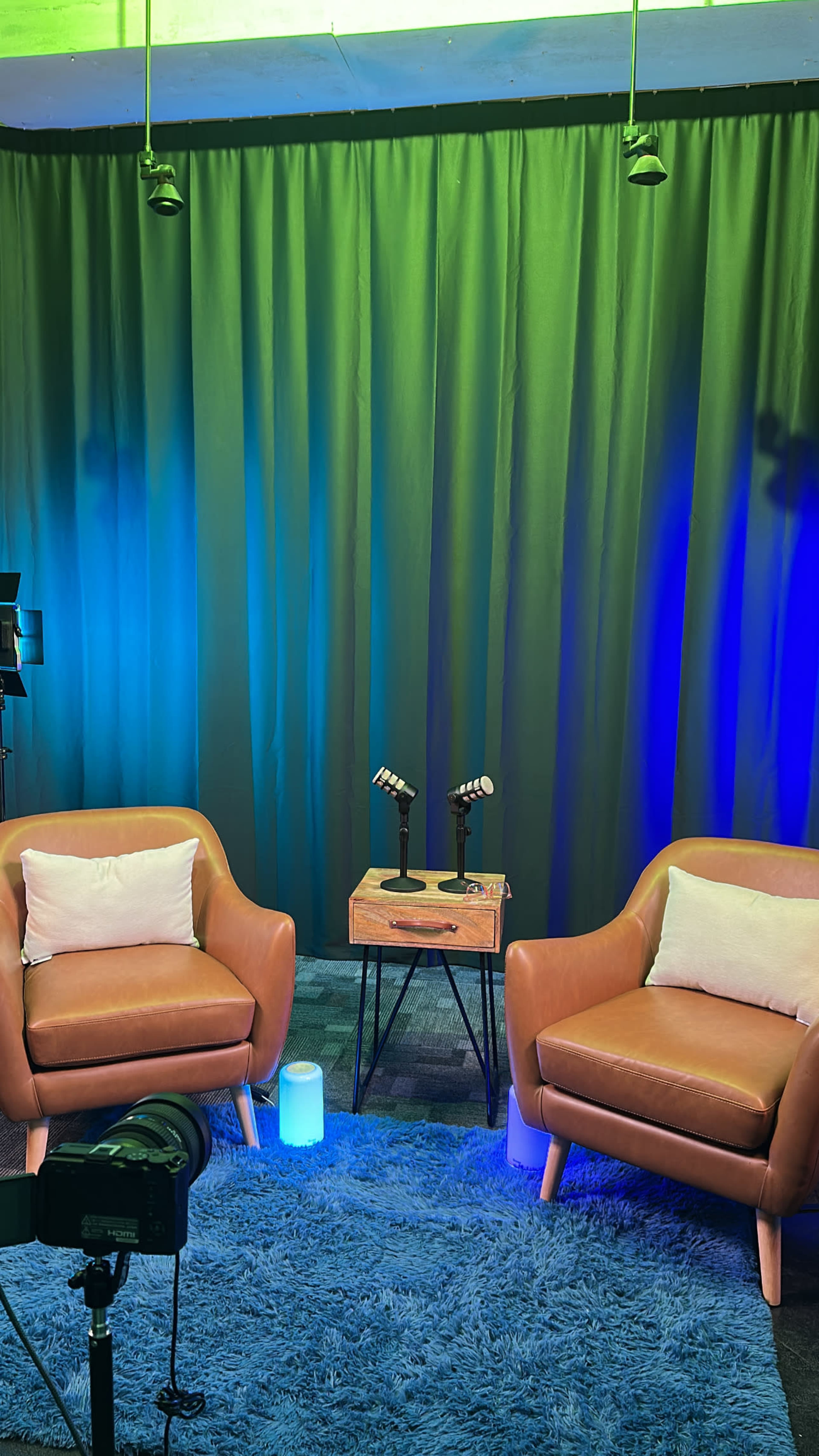 The image shows a studio setup with two brown chairs, a small wooden table with microphones, and blue and green lighting against a curtain backdrop.