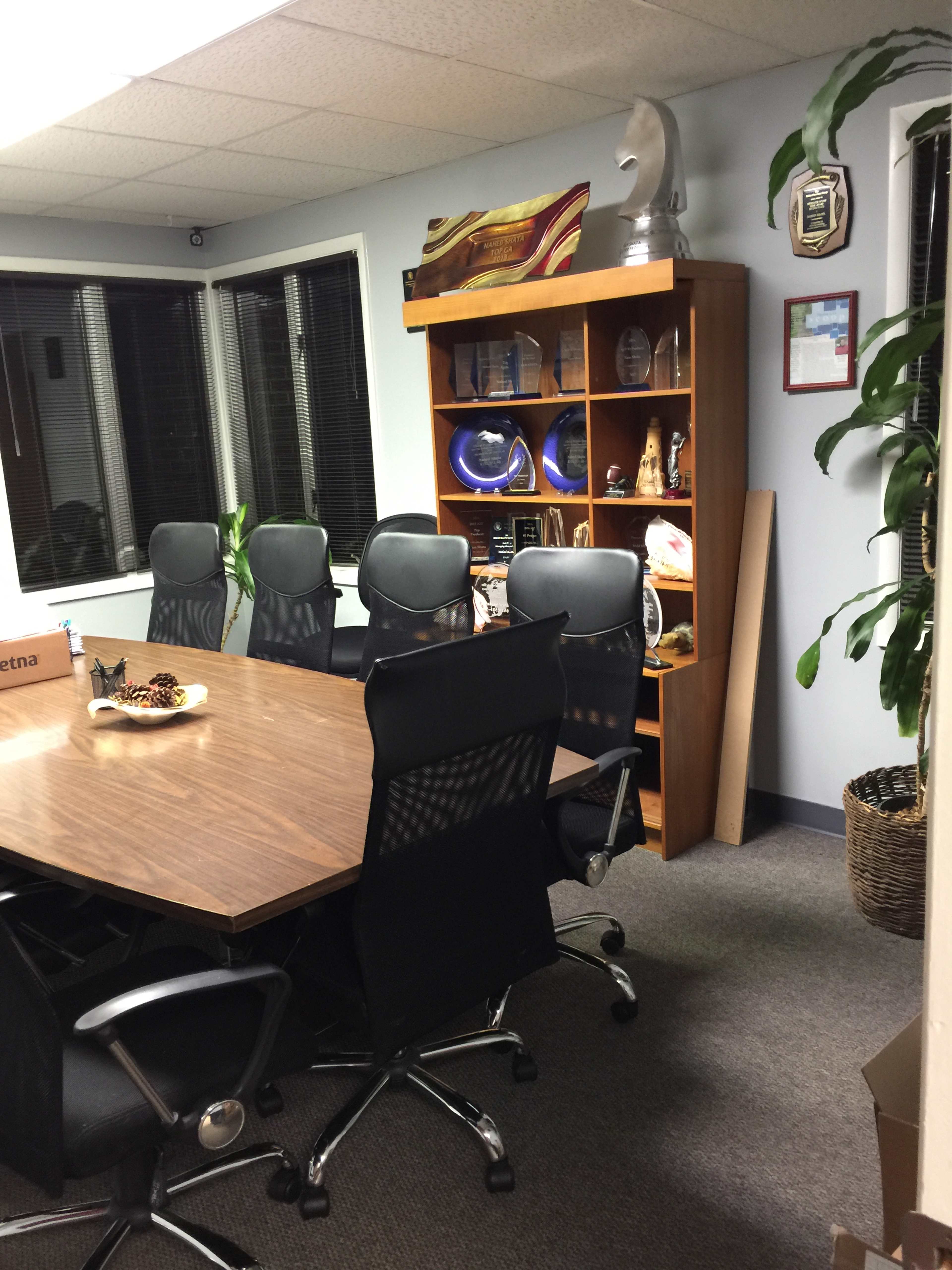 The image shows a conference room with a large wooden table surrounded by black office chairs, a bookshelf filled with decorative items and awards, and potted plants in the corners.