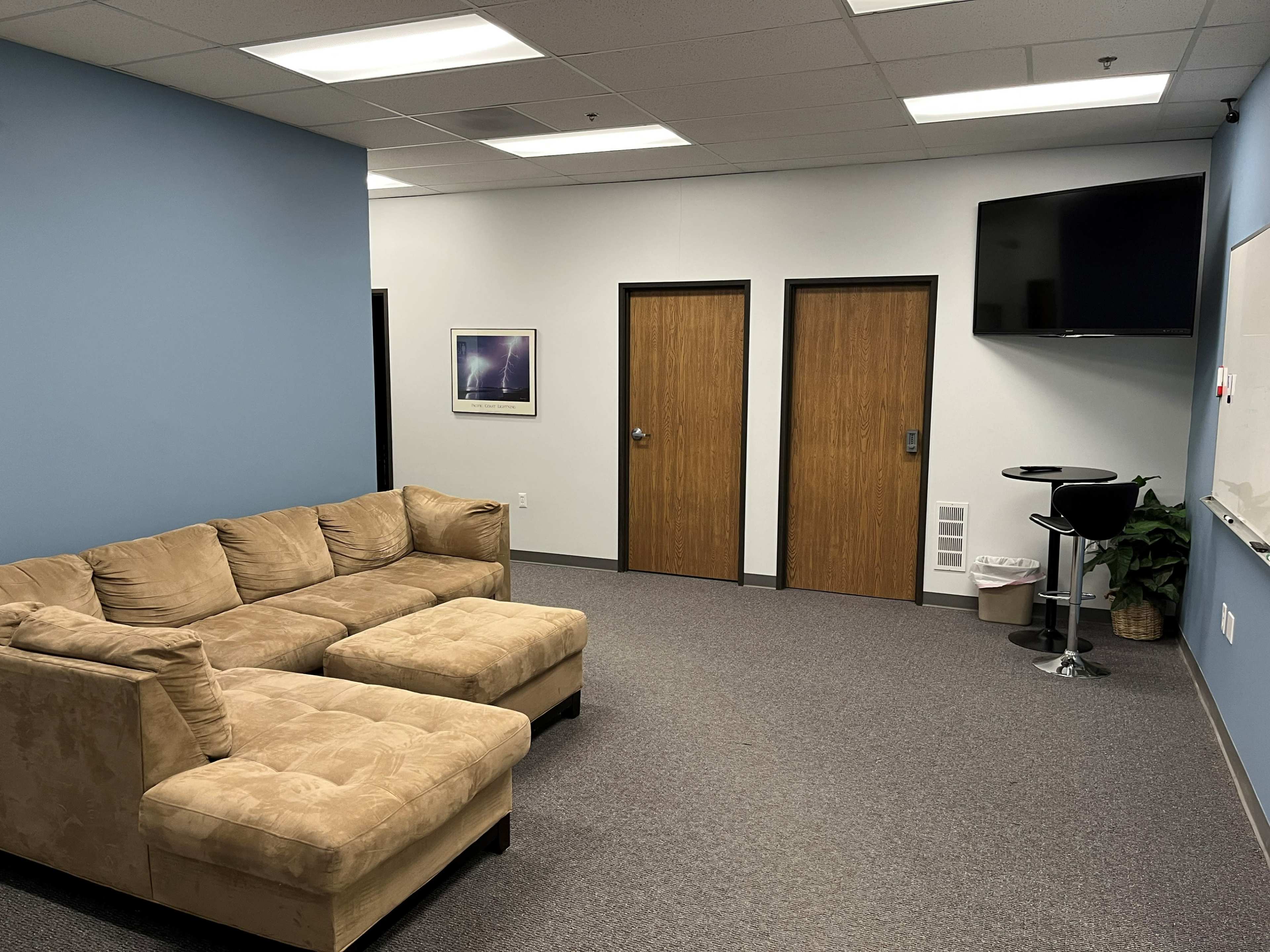 The room features a beige sectional couch, two closed wooden doors, a wall-mounted TV, and a small high table with a chair.