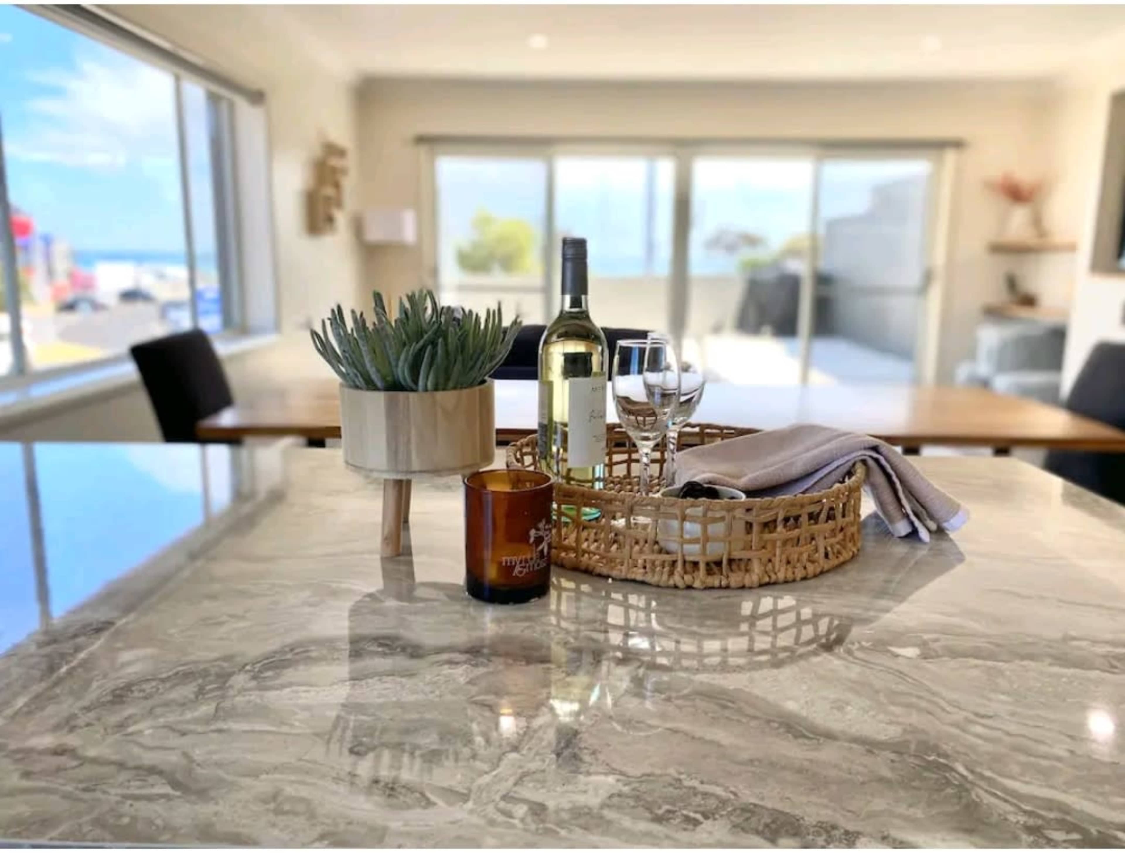 A bottle of white wine and two glasses are arranged on a woven tray with a small potted plant on a marble countertop, with a dining area visible in the background through large windows.