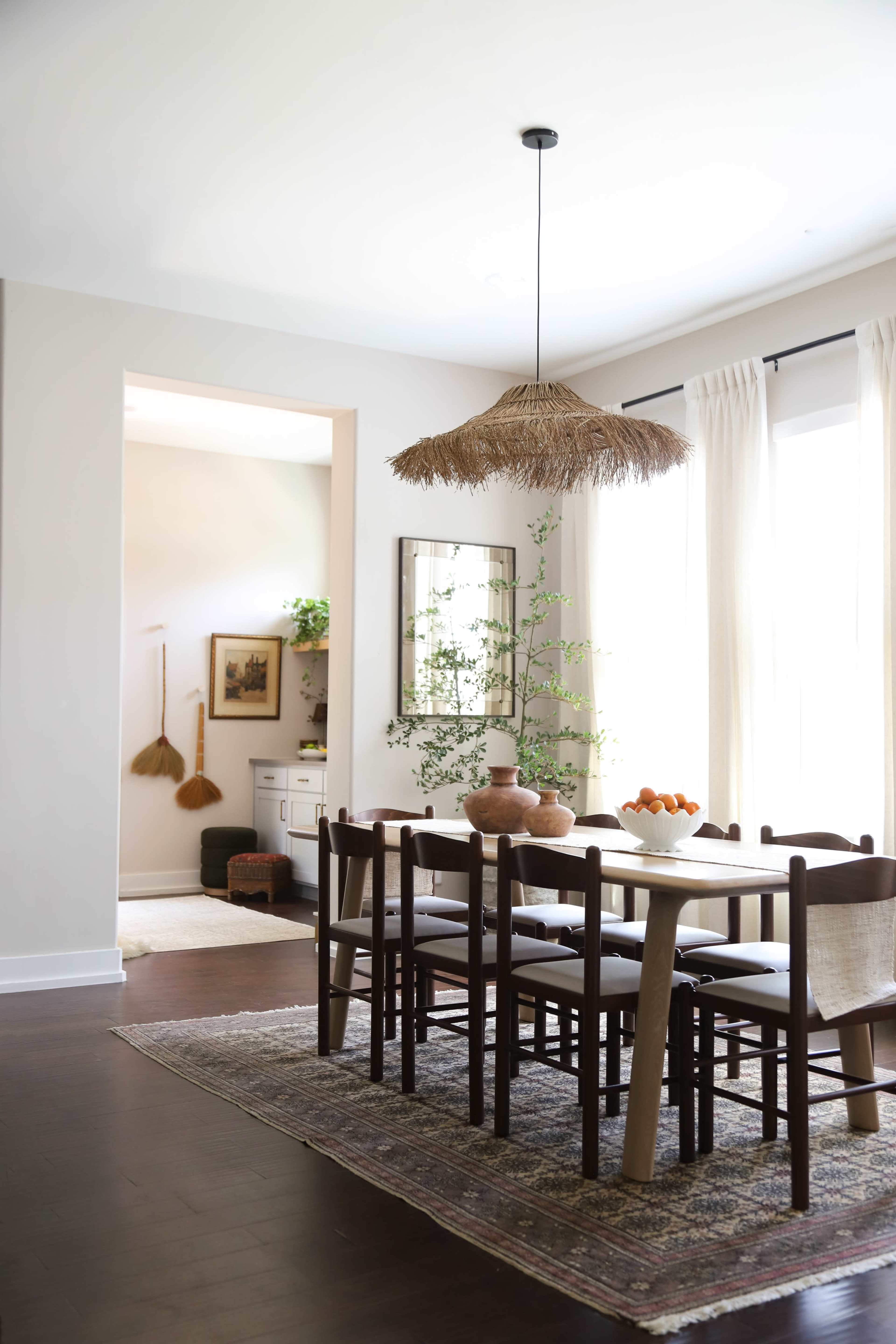 The image shows a dining room featuring a wooden table surrounded by chairs, a large straw light fixture, and a decorative arrangement of plants and fruits.