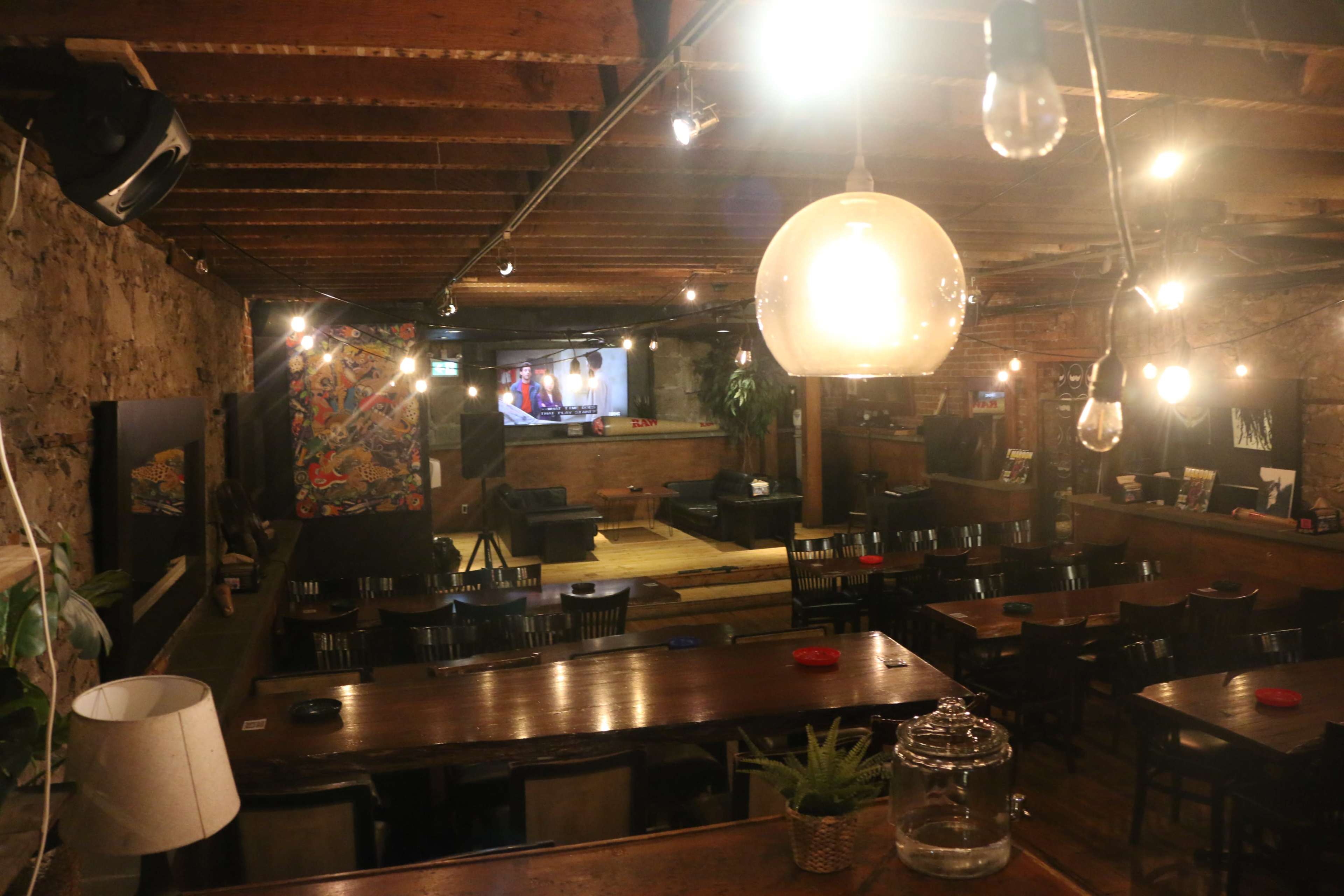A dimly lit interior of a restaurant or bar with wooden beams, several tables, and a large screen on the wall.