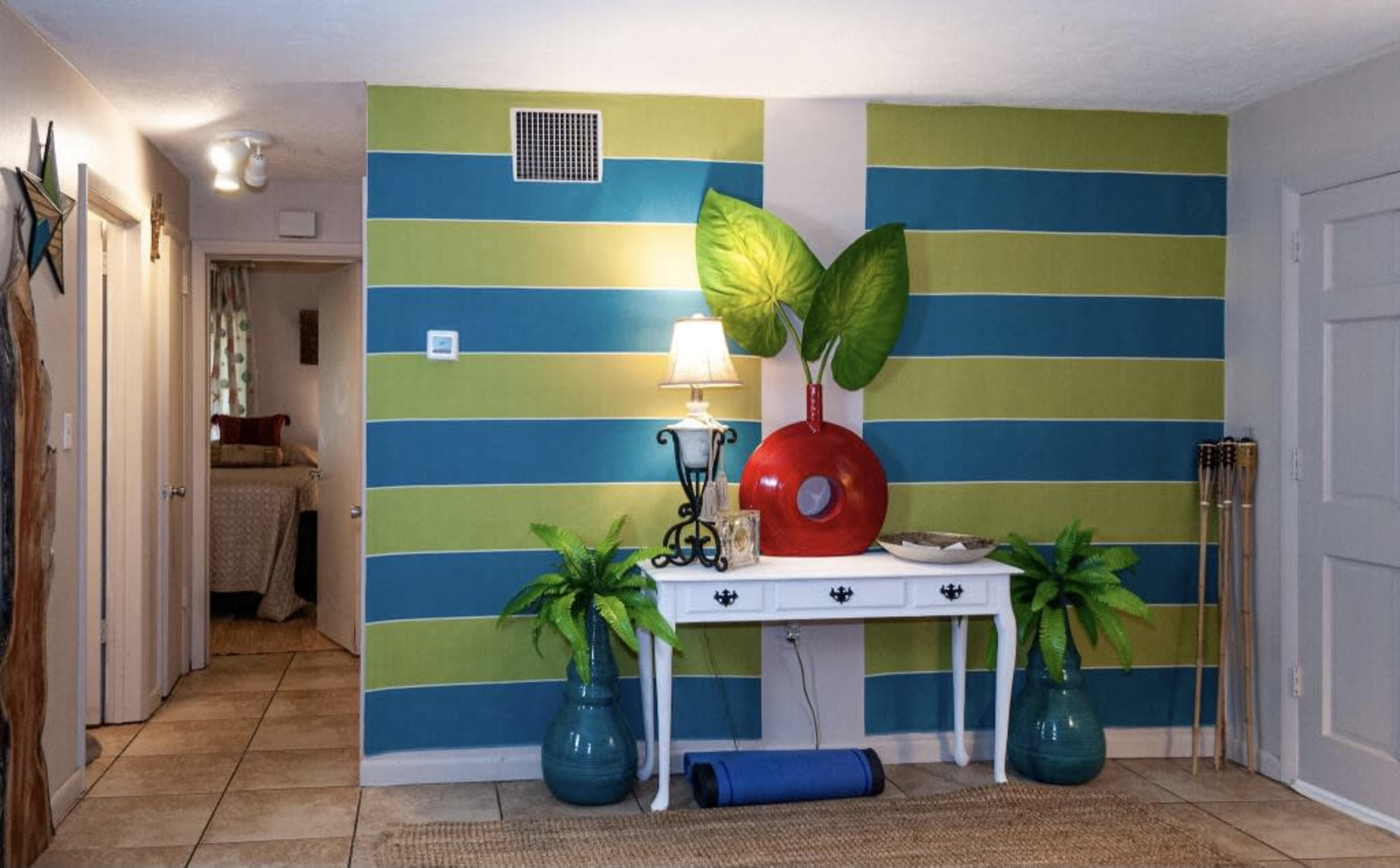 A brightly colored entryway featuring striped walls in blue and green, a white console table with a red decorative object and a lamp, and potted plants on either side.