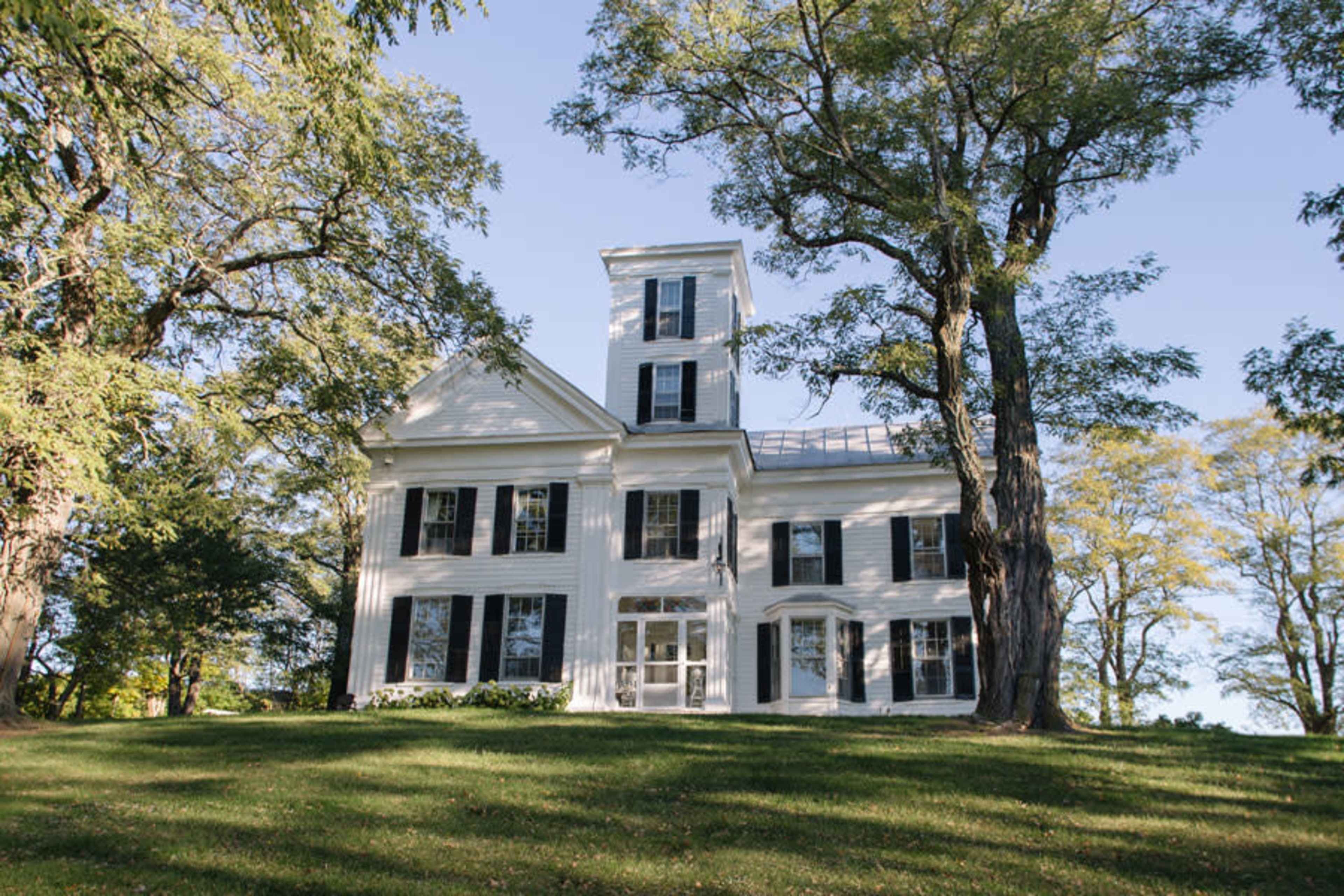 A large white house with black shutters stands on a grassy hill, surrounded by tall trees.