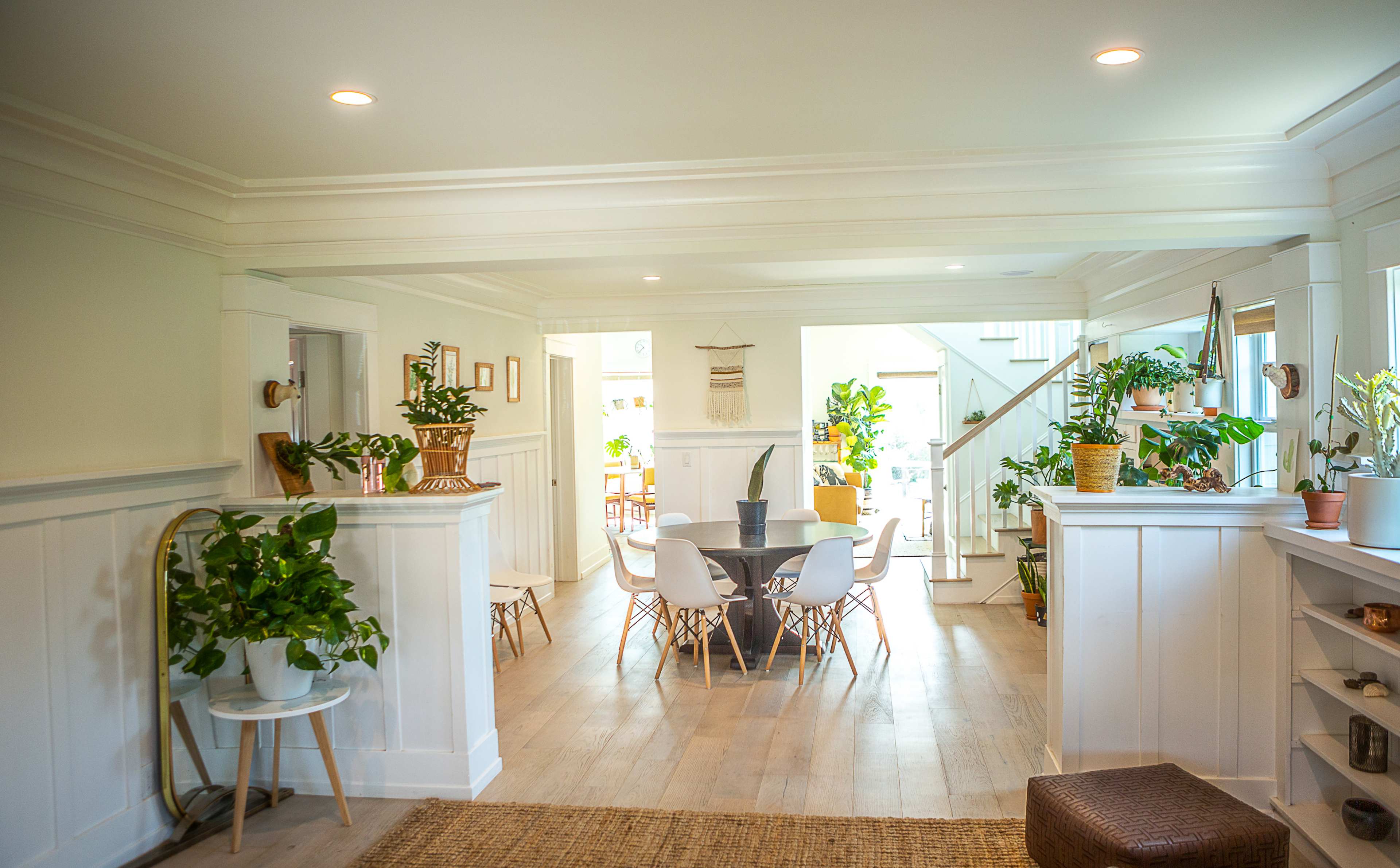 A bright and airy interior space with a dining area featuring a circular table surrounded by white chairs, potted plants, and a staircase in the background.