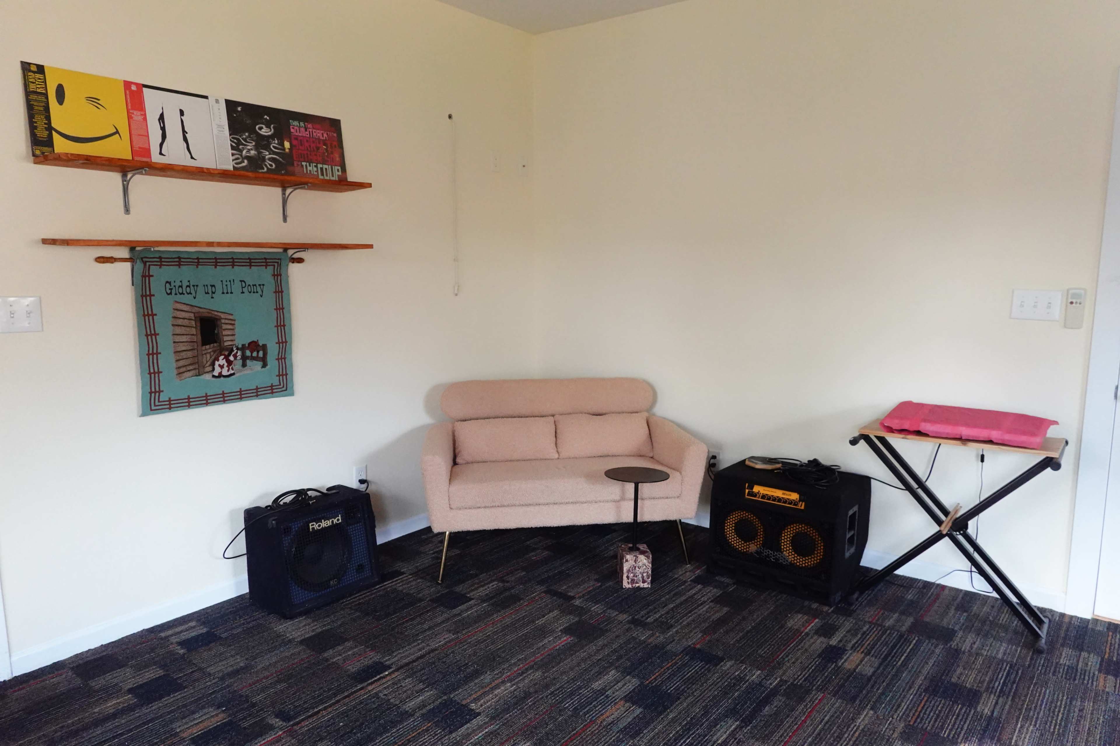 A small, modern living space features a pink couch, a round side table, and two speakers on either side, with a rack of vinyl records mounted on the wall.