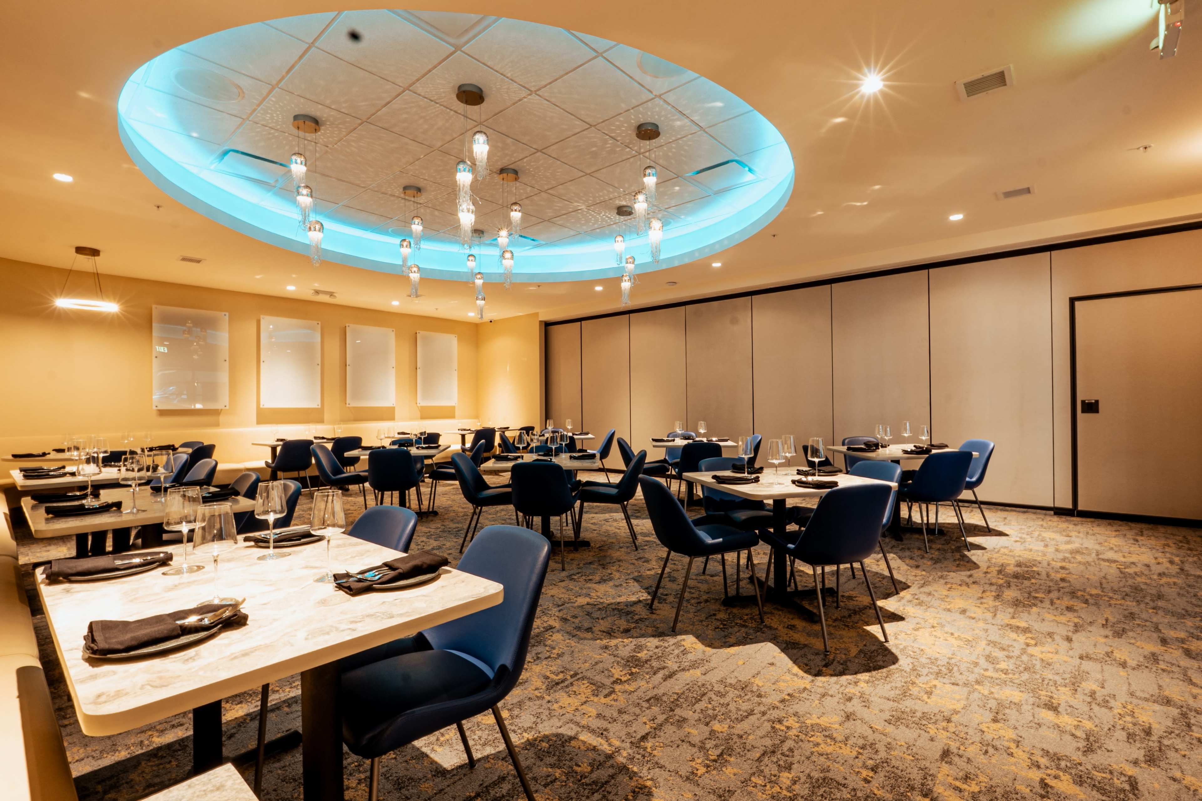 The image shows a modern restaurant interior featuring blue chairs, elegantly set tables, and a circular ceiling with lighting fixtures.