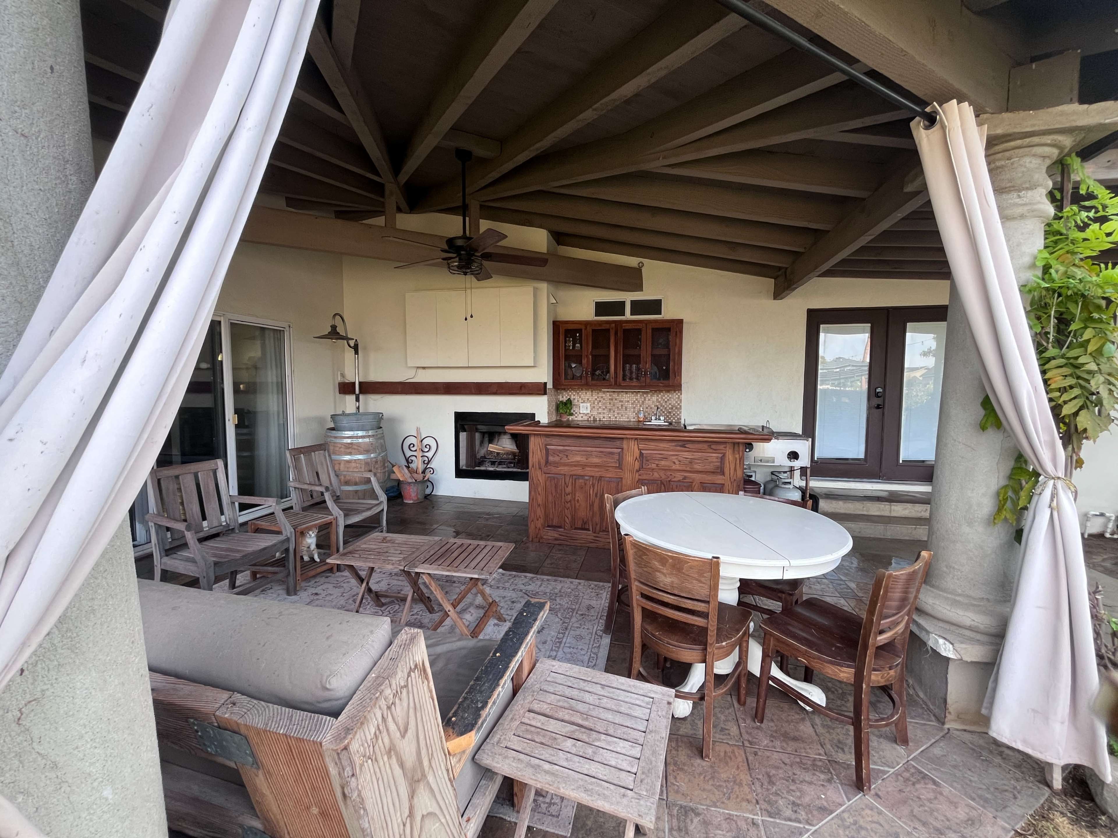 The image shows a spacious outdoor patio with wooden furniture, a bar area, and a cozy seating arrangement under a slanted roof.
