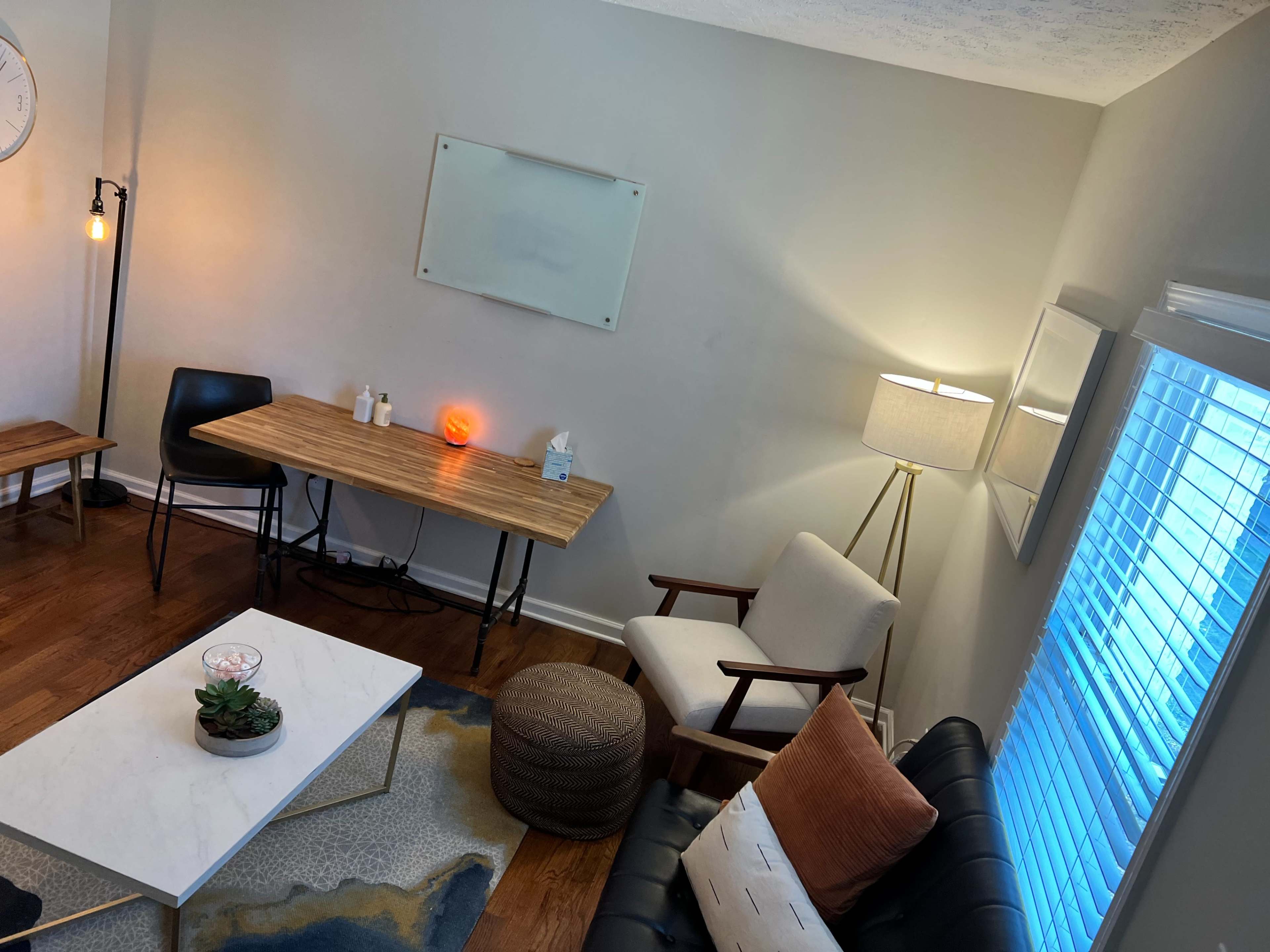 The image shows a cozy living space with a wooden table, a black chair, a couch, and decorative elements like a lamp and a small plant on a coffee table.
