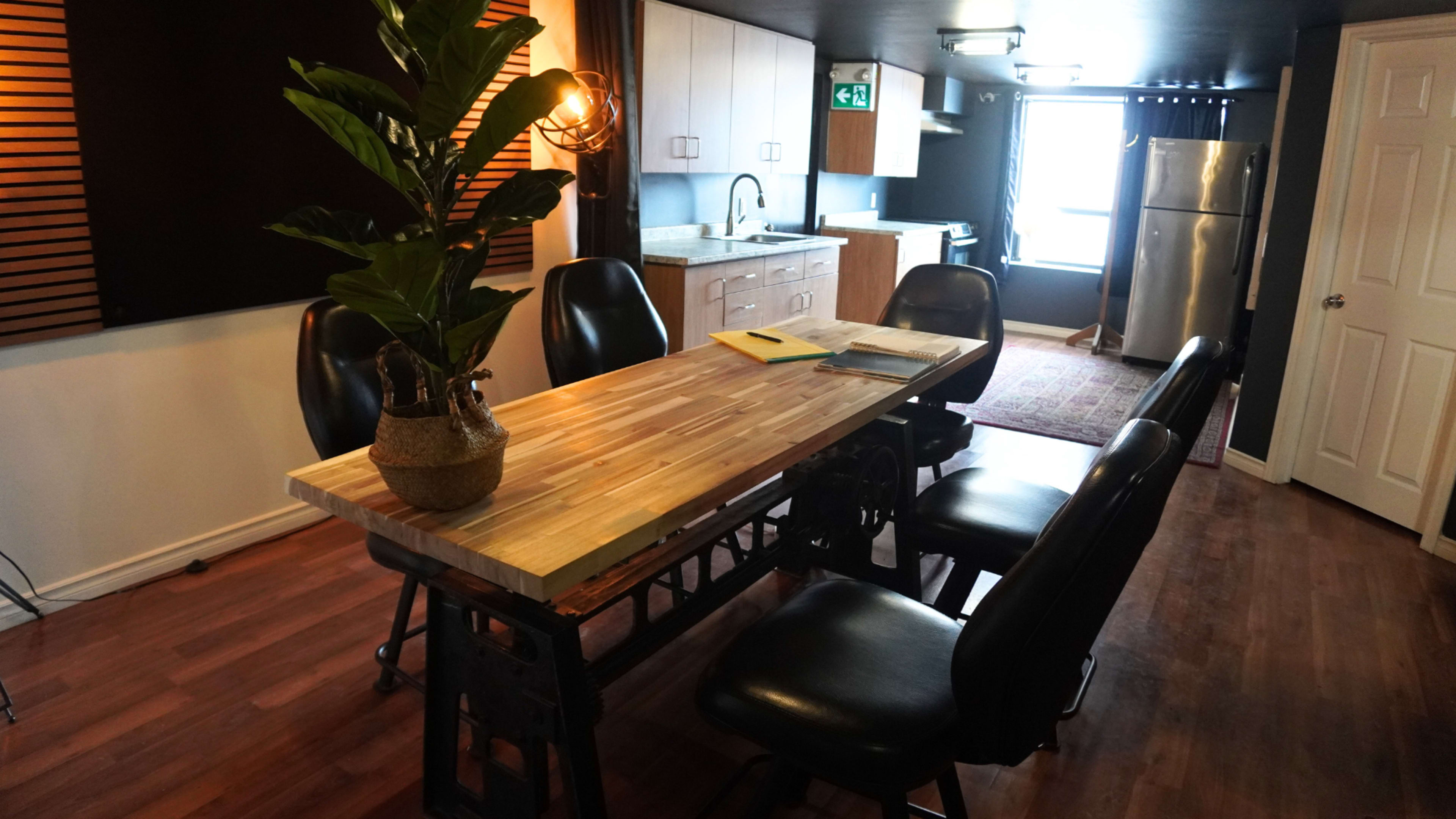 A modern kitchen area features a wooden dining table with black chairs, a decorative plant, and a kitchenette in the background.