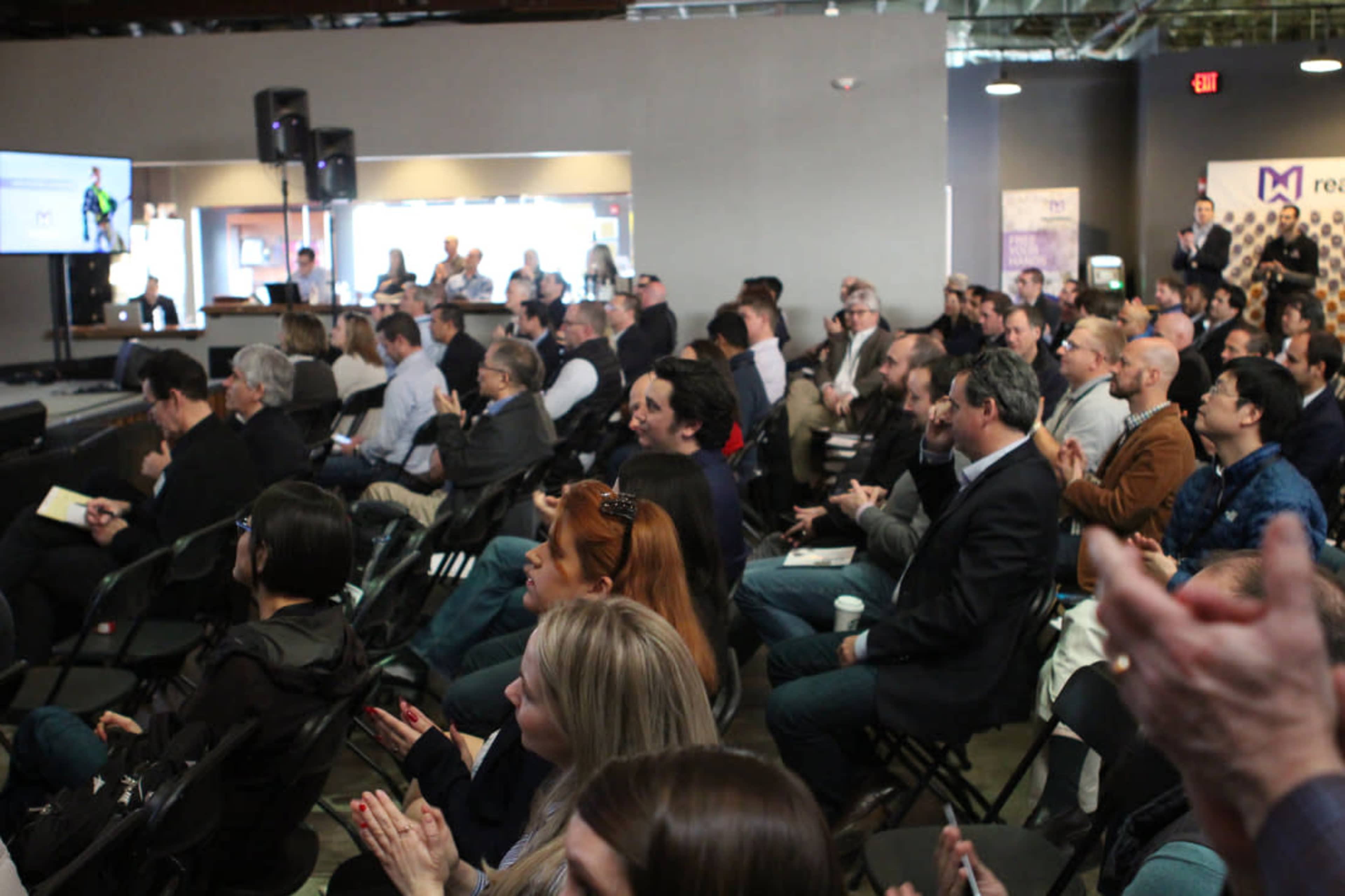 A diverse audience sits in a conference room, attentively watching a presentation on a large screen.