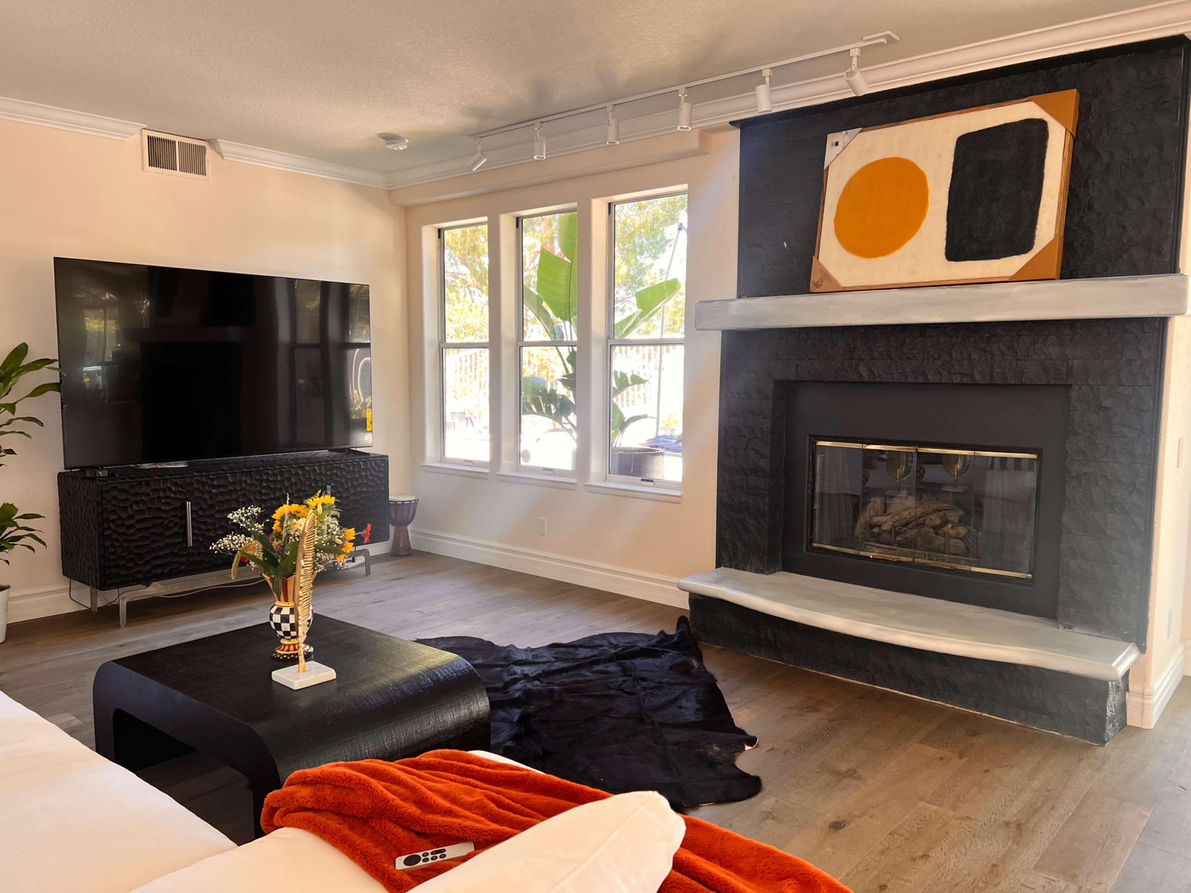 The living room features a large TV mounted on the wall, a modern fireplace with a white ledge, and a coffee table, surrounded by large windows and greenery.