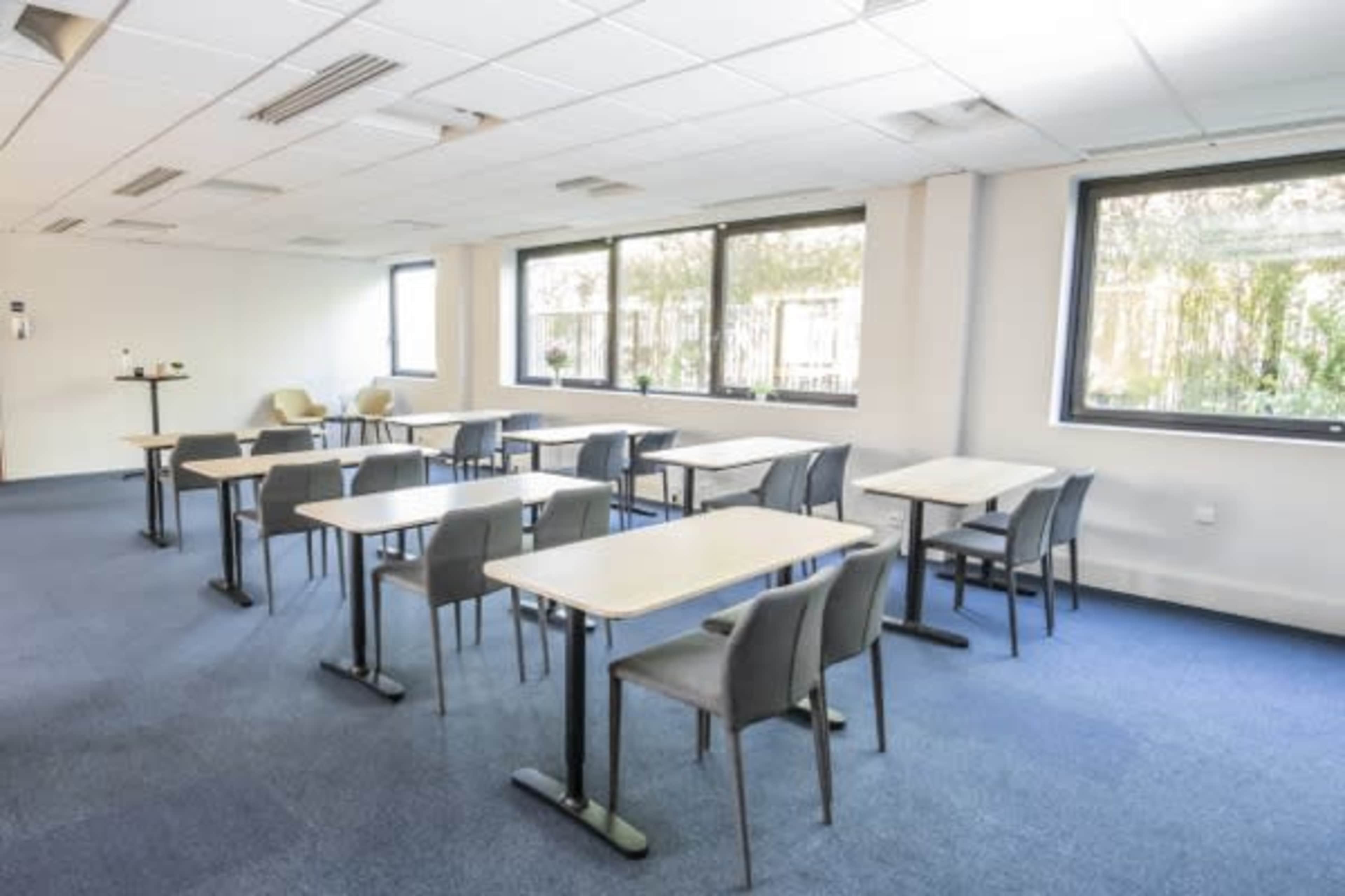 A room with several tables and chairs arranged in a classroom layout, featuring large windows with greenery outside.