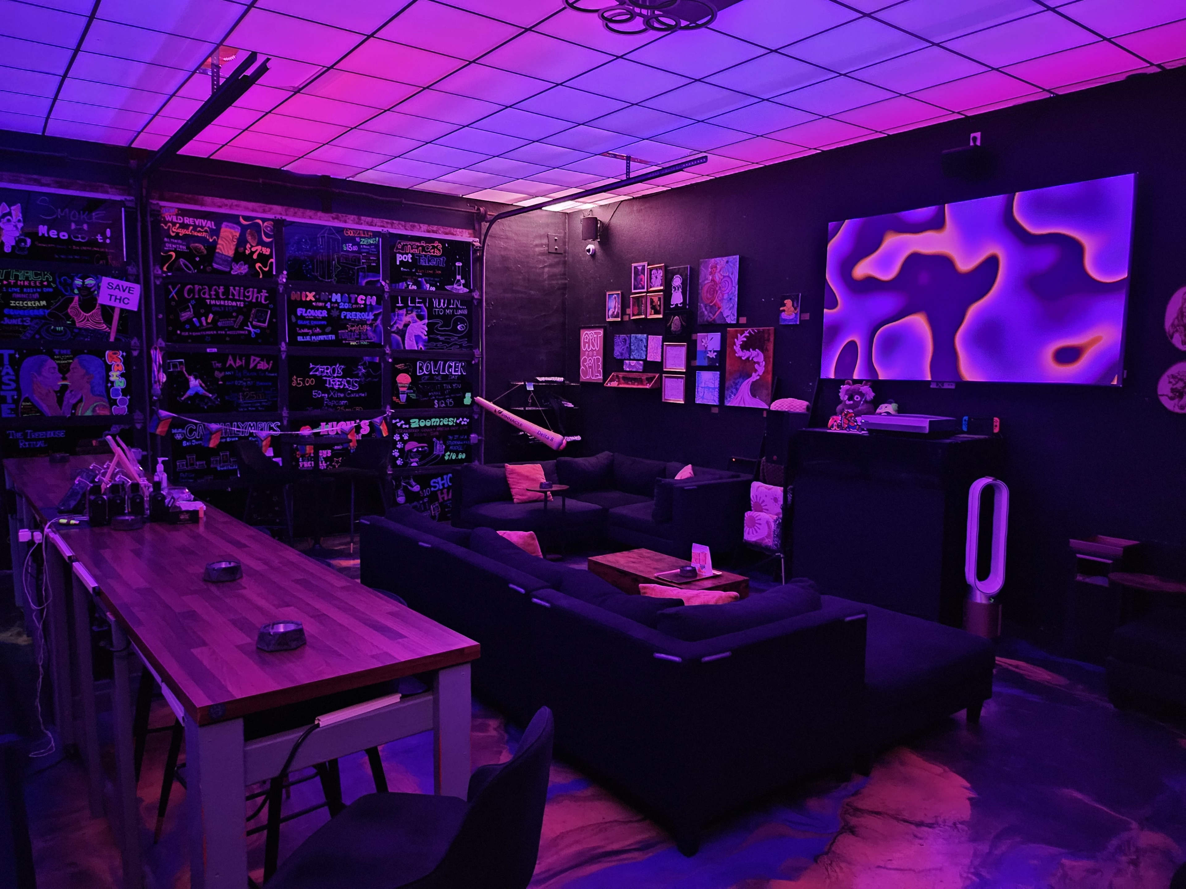 The image shows a modern lounge area with black walls, vibrant purple lighting, and comfortable couches arranged around a wooden table.