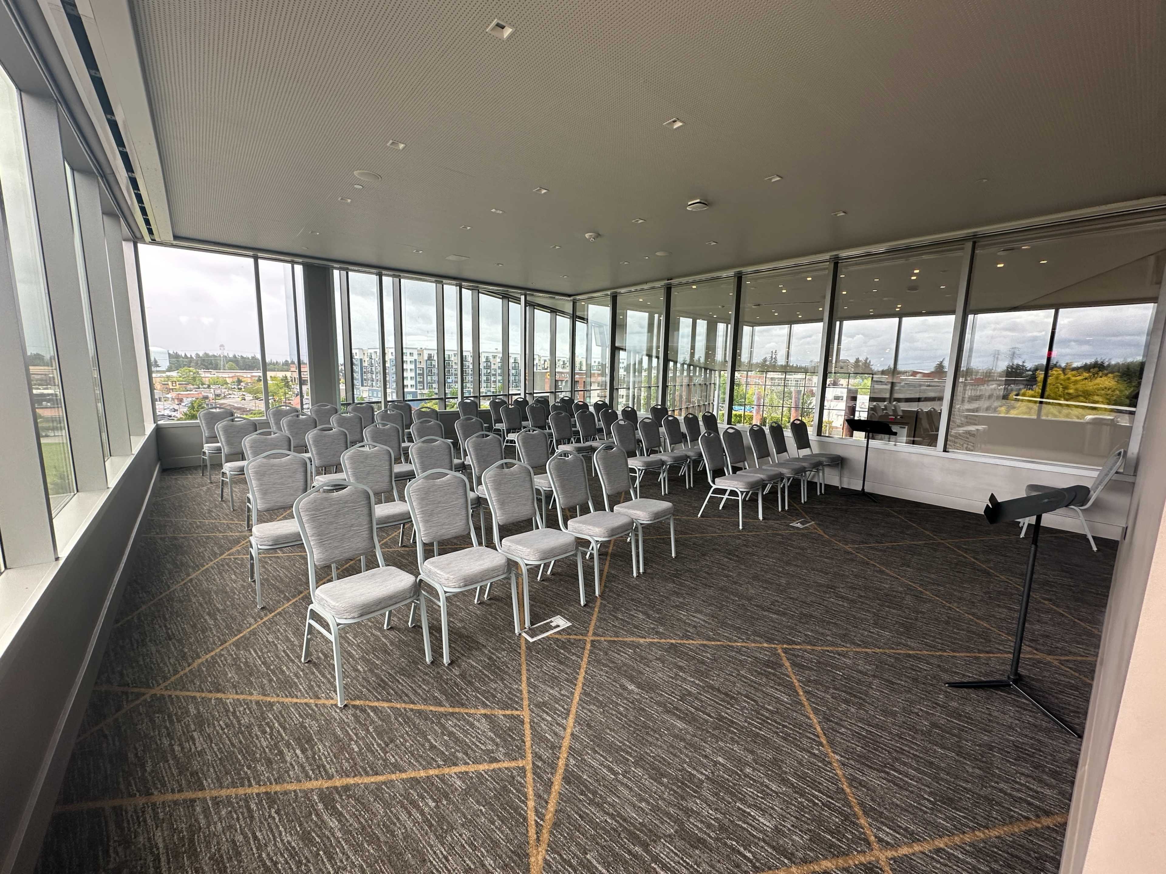 The image shows a conference room with rows of gray chairs arranged facing a front area with large windows overlooking an outdoor landscape.