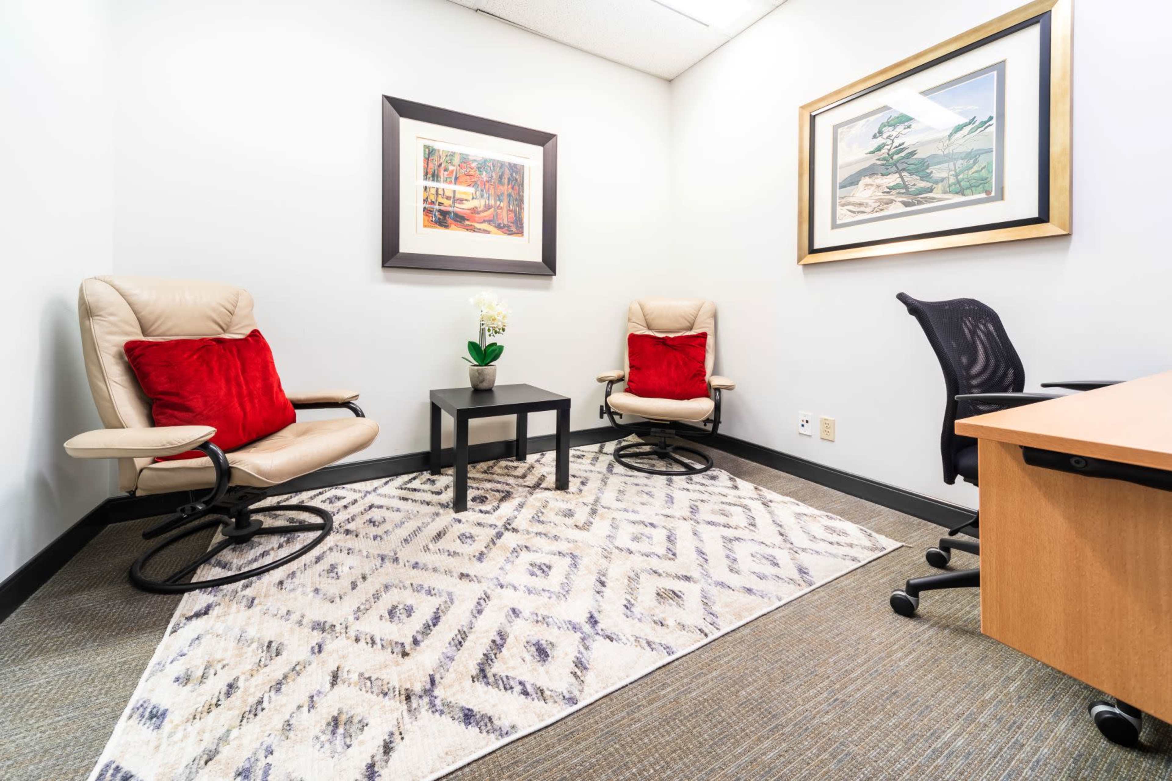 A small office space with two chairs, a coffee table, a plant, and a desk against the wall, decorated with framed artwork.