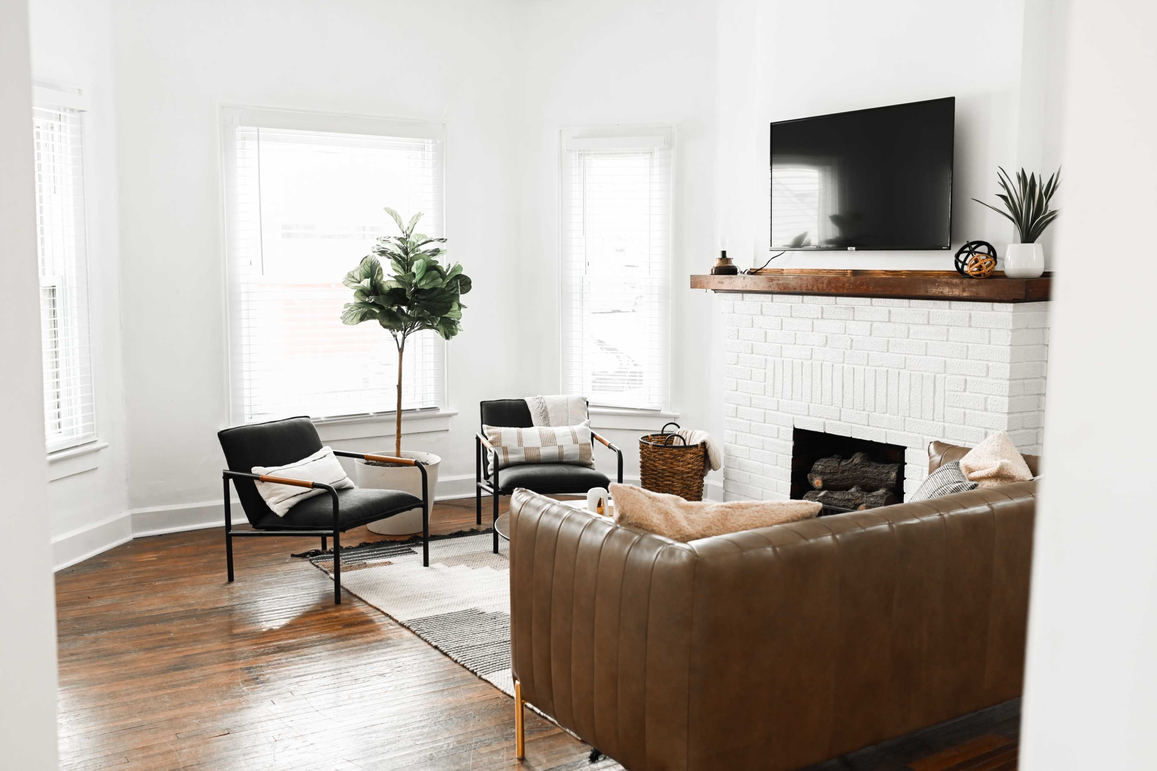 A modern living room features a brown leather sofa, two black chairs, a potted plant, and a fireplace with a television mounted above it, all set against white walls and wooden floors.