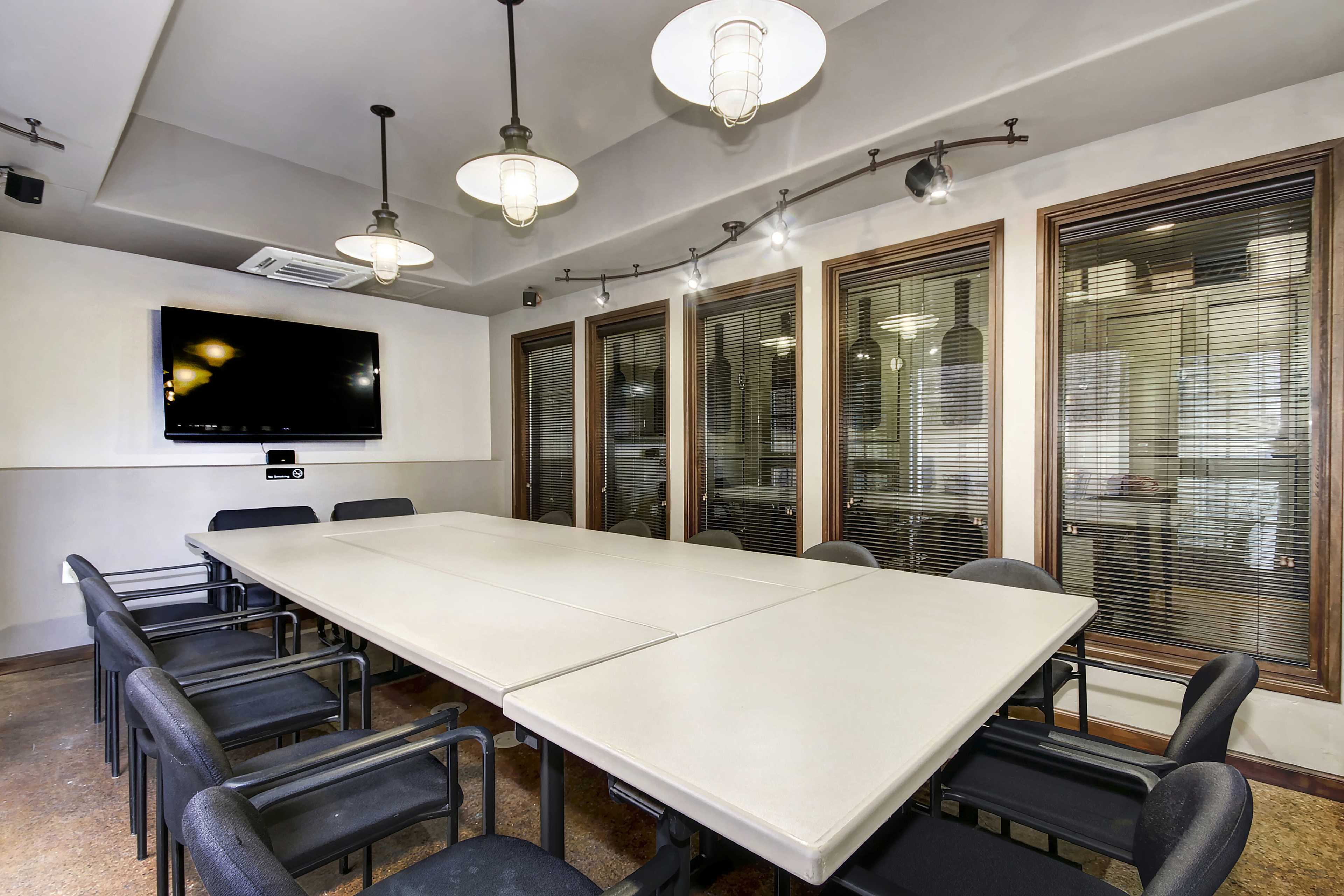 A conference room features a long table surrounded by chairs, with a television mounted on the wall and large windows with blinds.