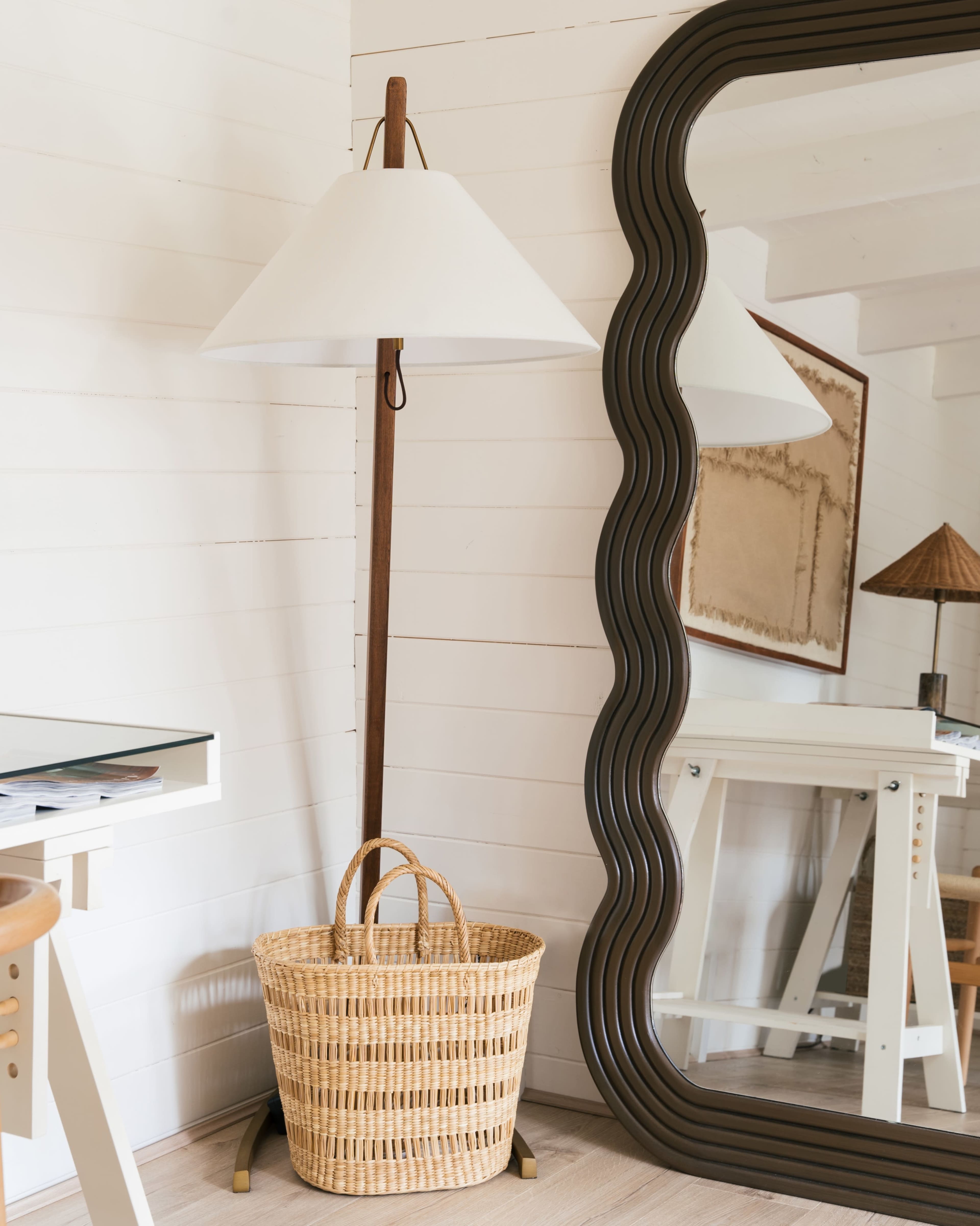 A floor lamp with a white shade next to a woven basket, positioned near a wavy-edged mirror on a white wooden wall.