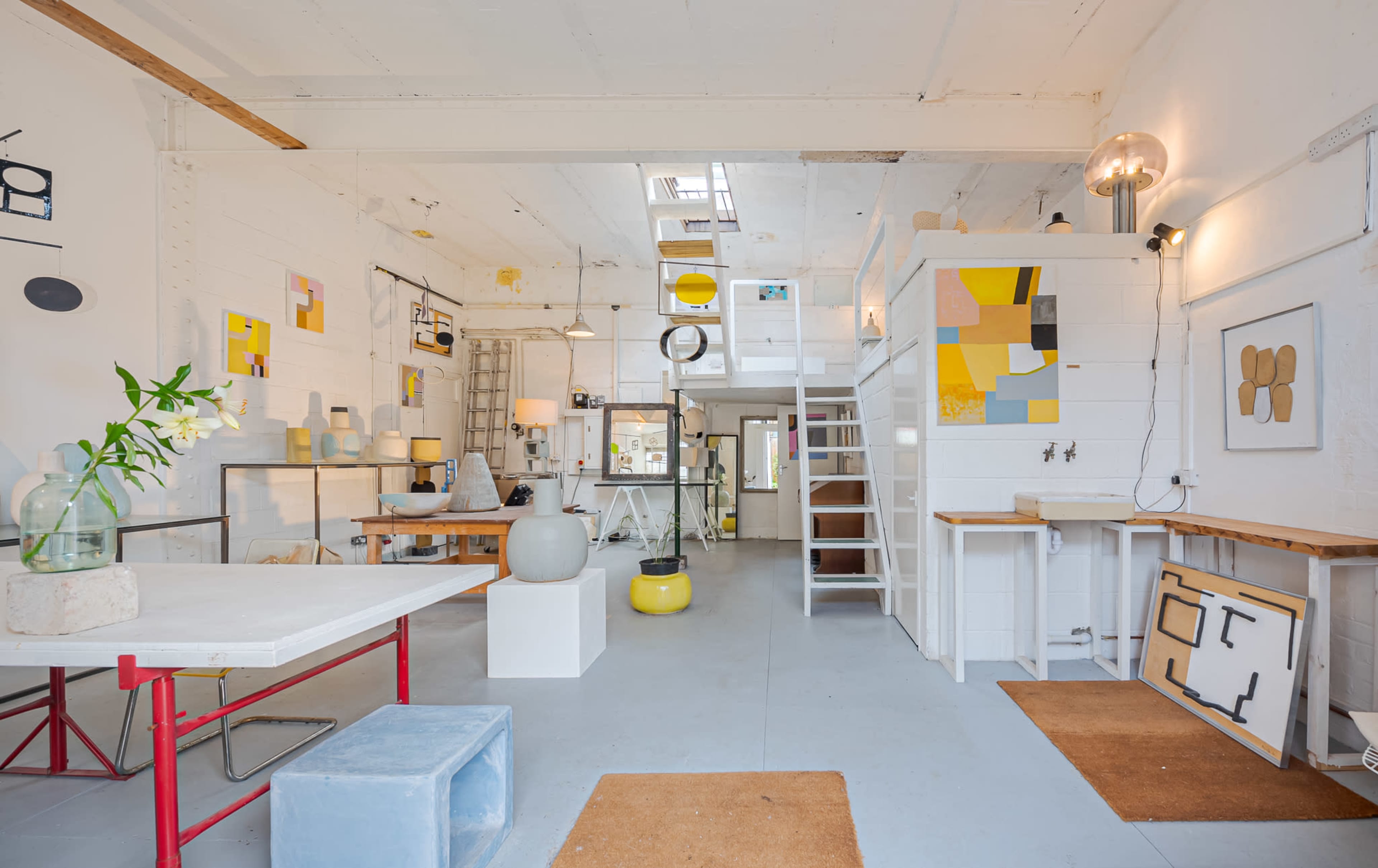 A spacious, well-lit studio with various artworks displayed on the walls and a mix of furniture, including tables, stools, and decorative items.
