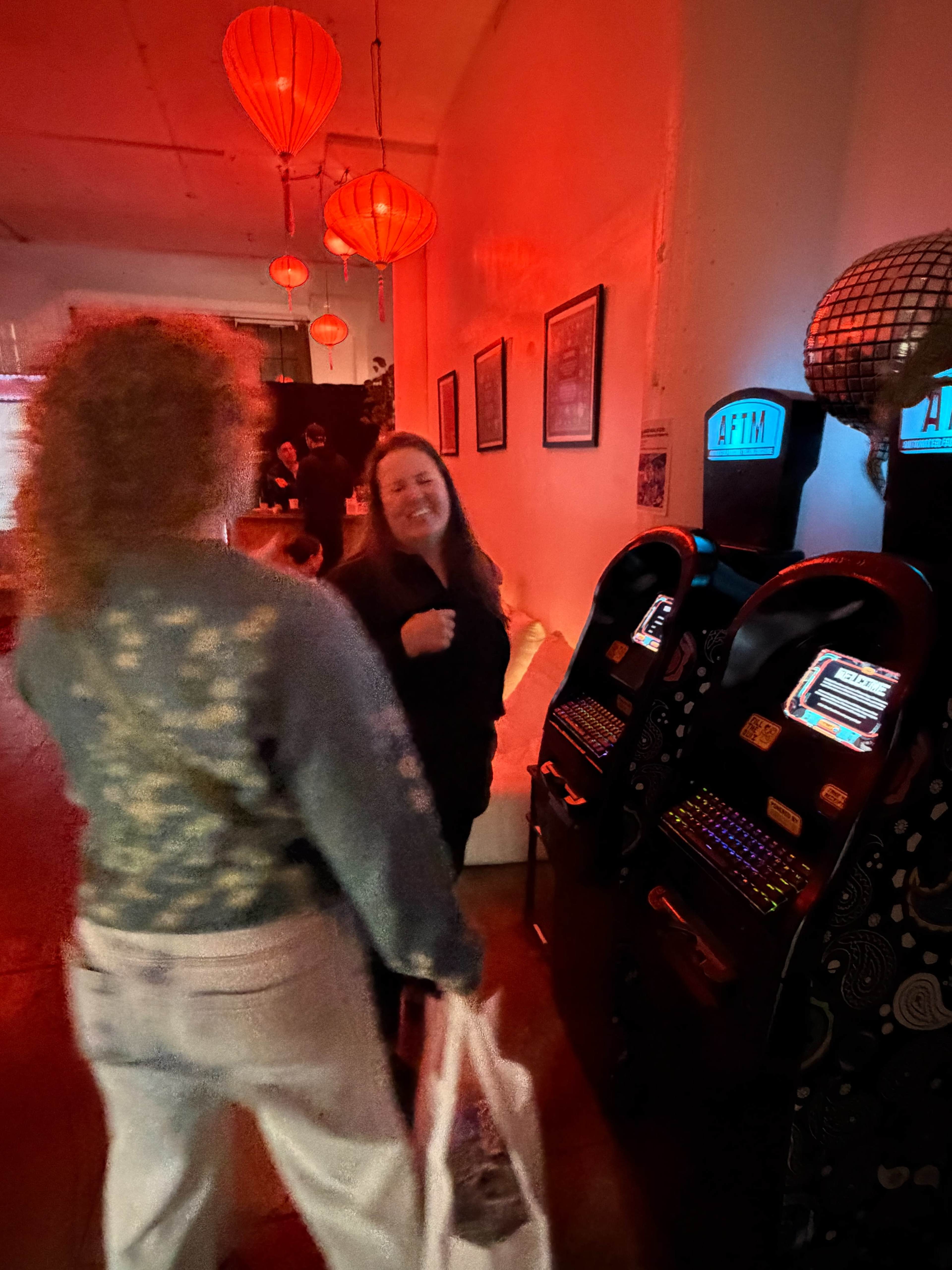 The image shows two women interacting in a venue with red lighting and two vintage arcade machines in the background.