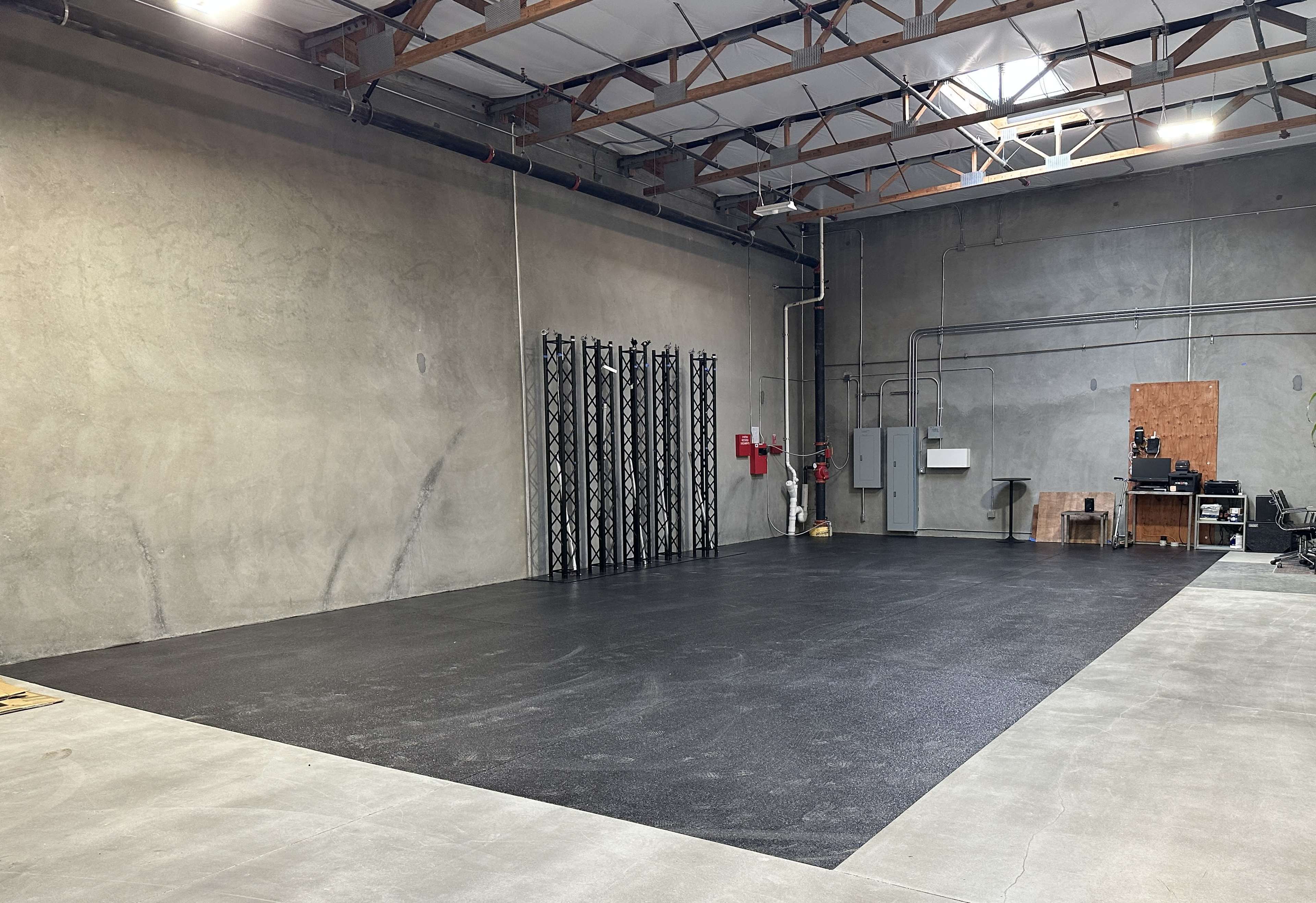 The image shows a large, empty industrial warehouse space with concrete walls and a dark rubber floor, featuring a fire extinguisher and electrical panels along one side.