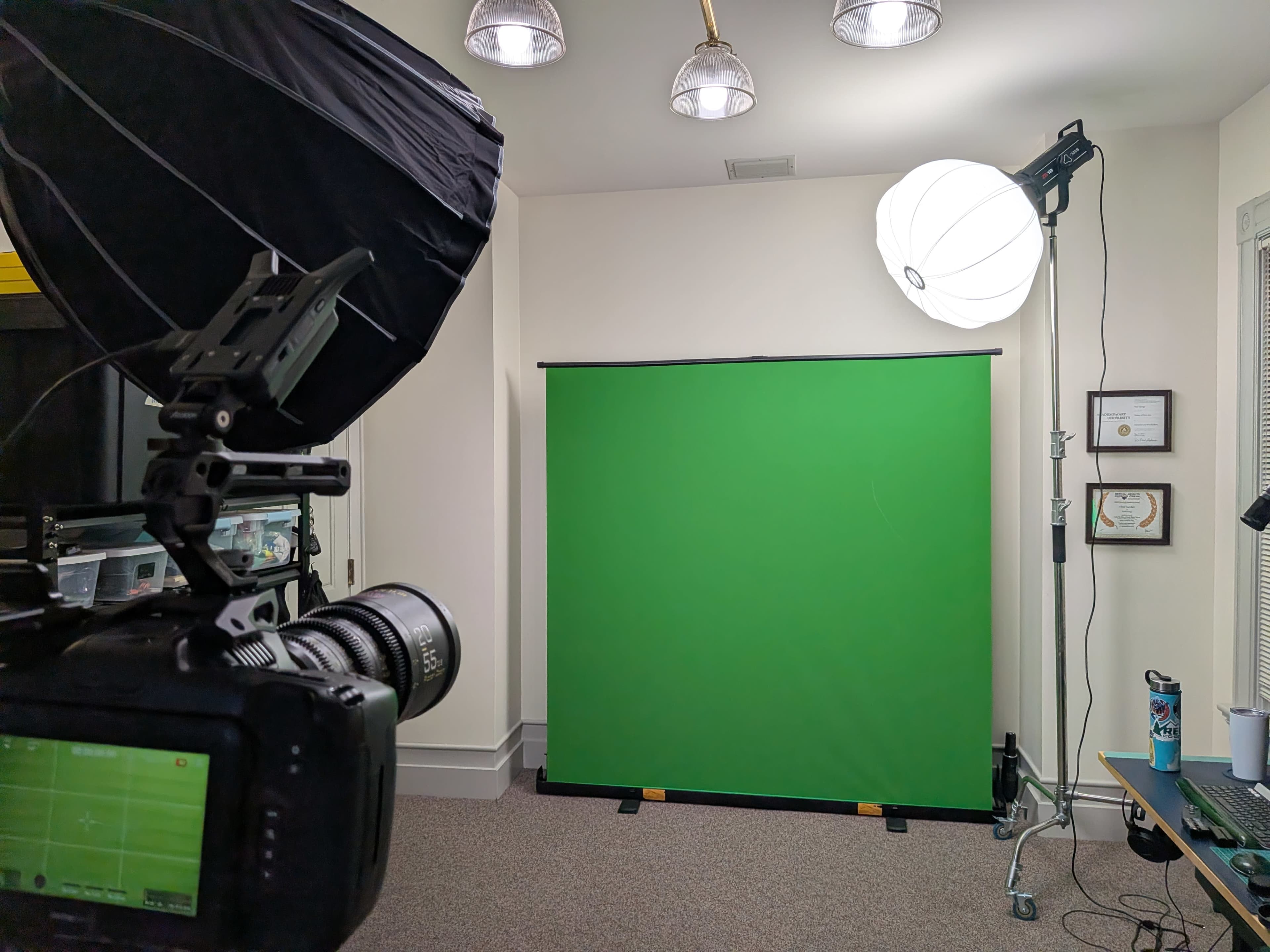A camera is positioned to capture a room featuring a green screen, a large softbox light, and a desk with various items.