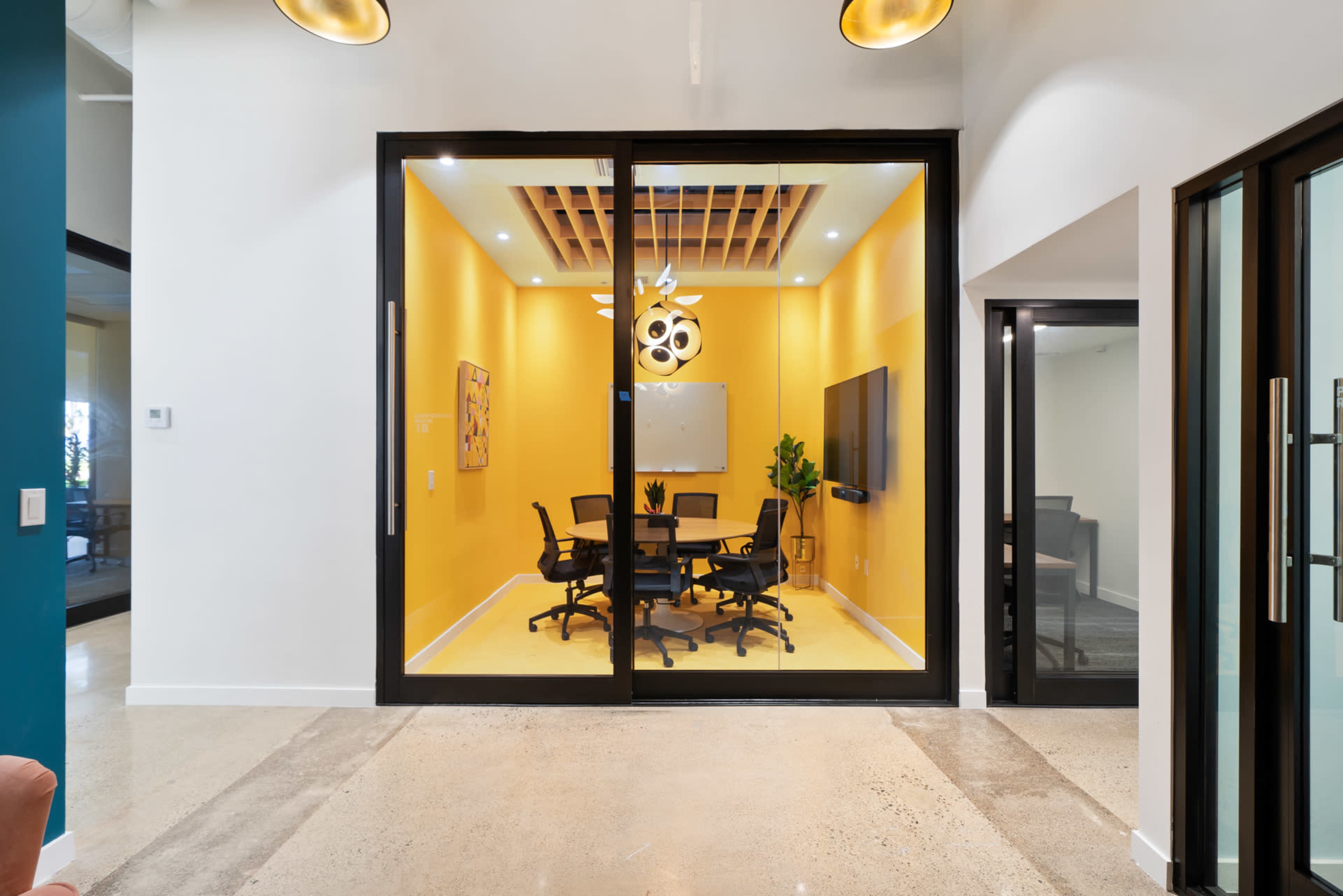 A conference room with a yellow wall, a round table, and four black chairs is visible through a large glass window.