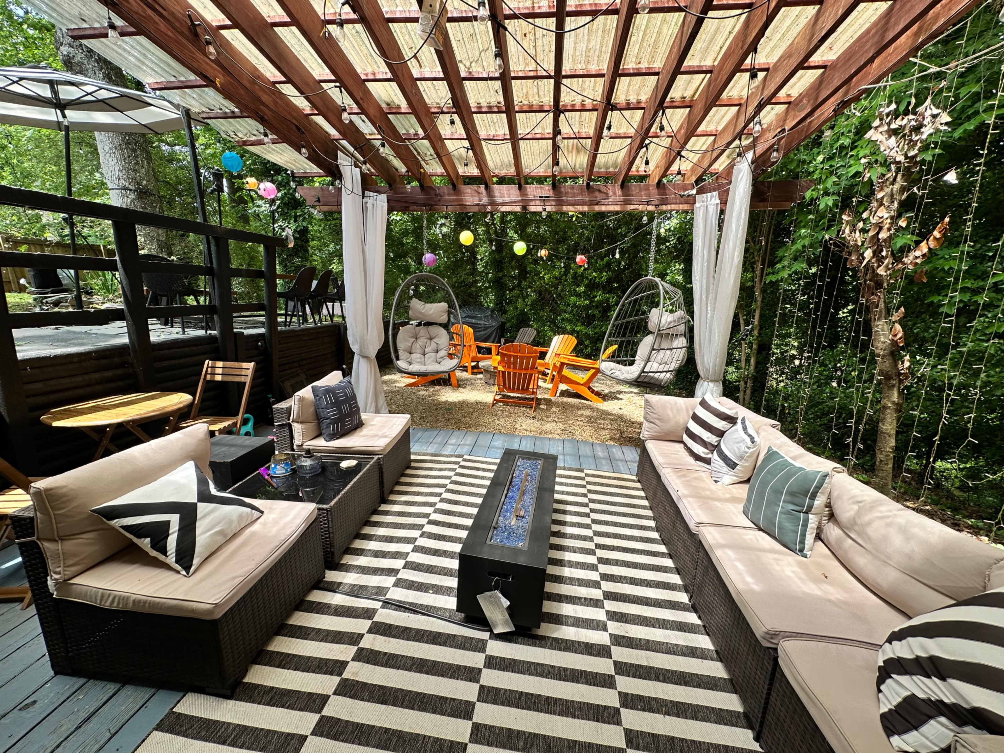 A covered outdoor patio with sectional seating, a fire pit, hanging chairs, and colorful decorations surrounded by greenery.