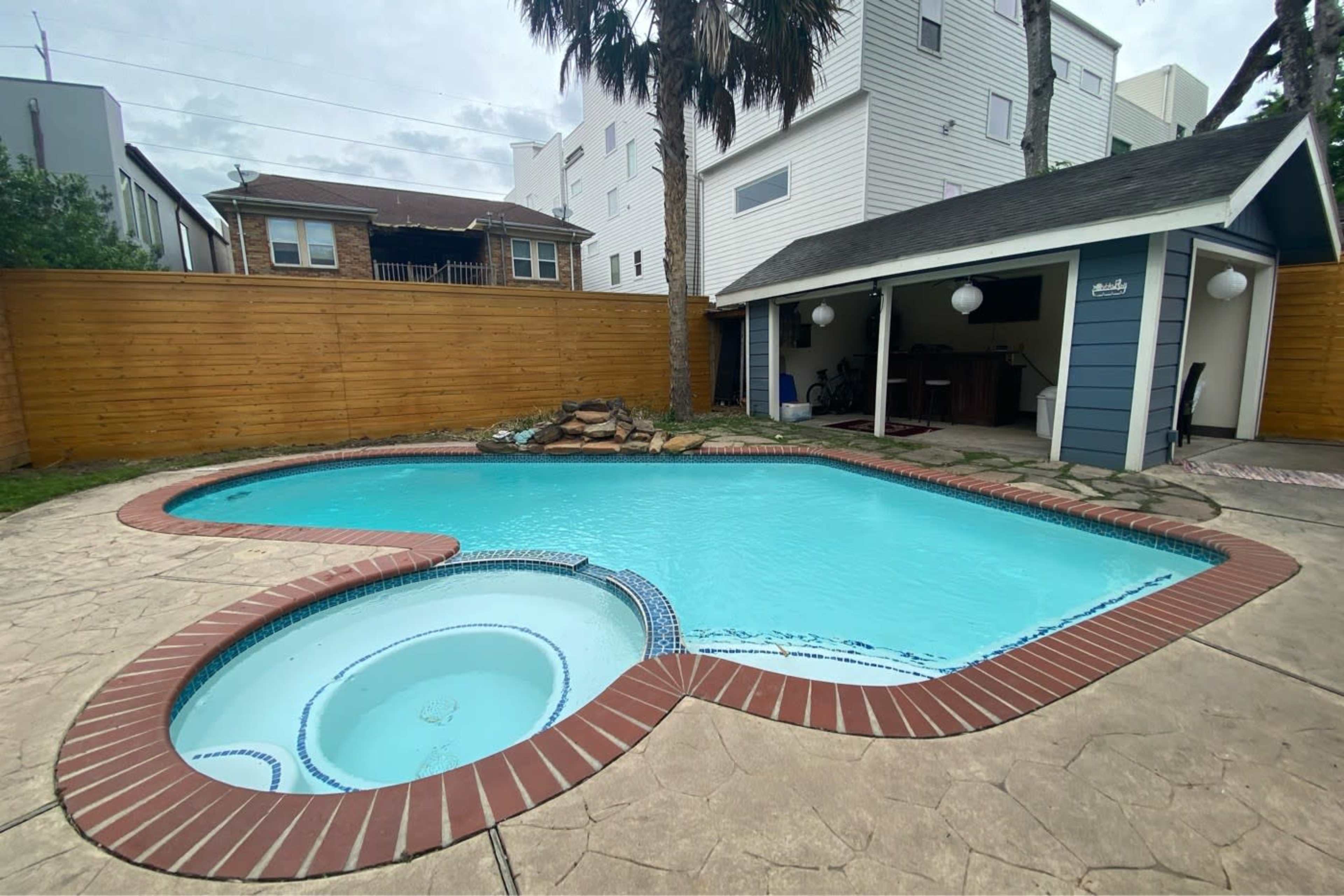 A turquoise-shaped pool surrounded by a stone patio and a wooden fence, with a small outdoor structure in the background.